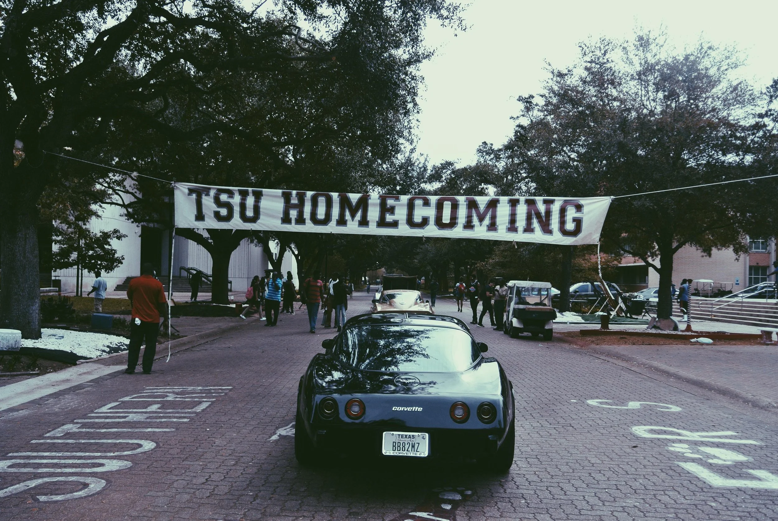 A black Chevrolet Corvette parked in front of a banner that reads 'TSU HOMECOMING'. The setting appears to be a university campus with trees, people, and vehicles, indicating a homecoming event.