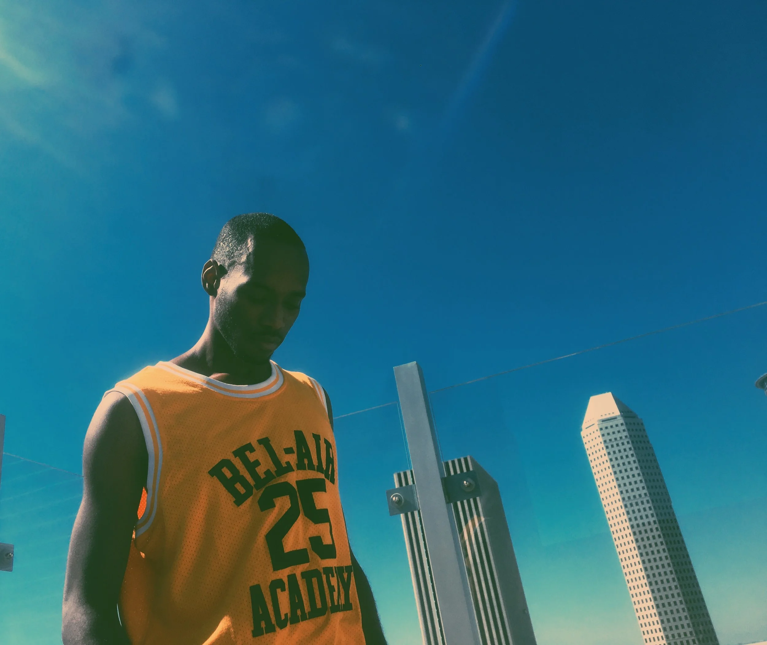 Young man in yellow basketball jersey with number 25 and 'Bel-Aire Academy' written on it, standing outdoors with city buildings in the background under a clear blue sky.