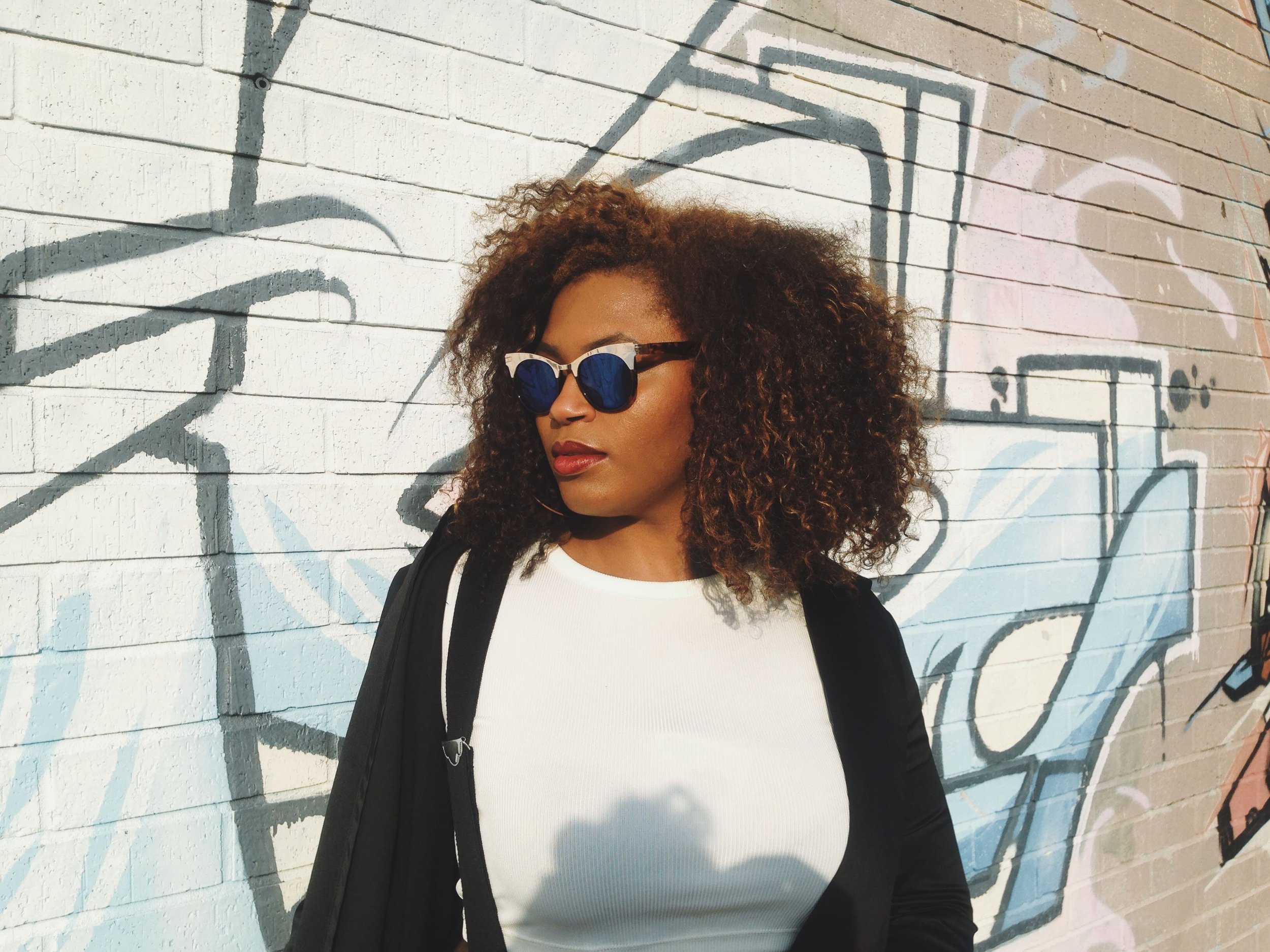 A woman with curly hair wearing sunglasses, a white top, and a black jacket is standing in front of a graffiti wall.
