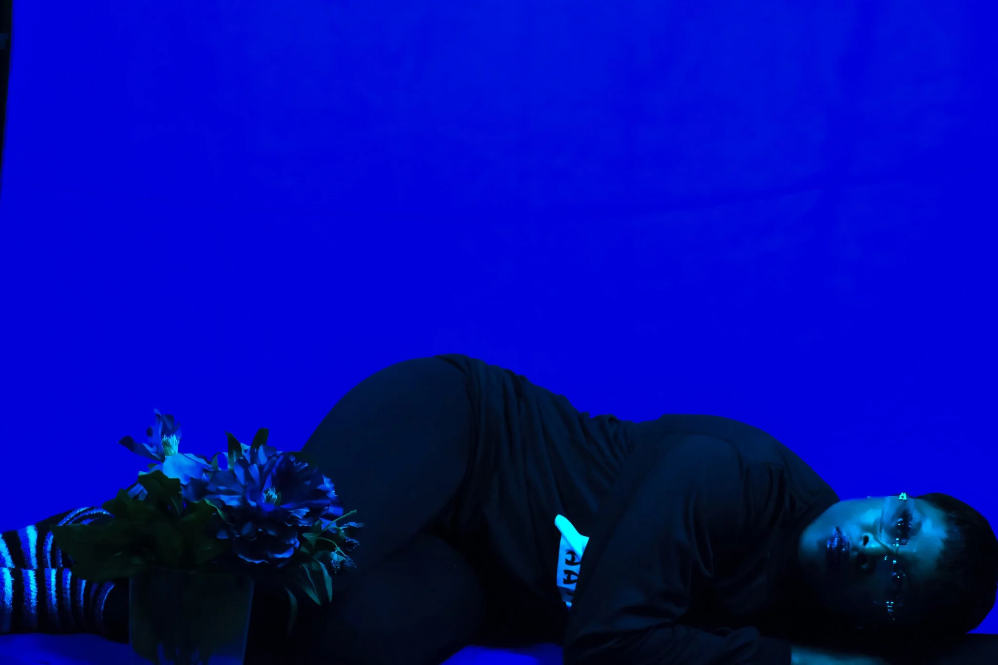 Person with glasses and short hair lying on the floor with blue background, near a vase of blue and white flowers, dressed in black shirt, black pants, and striped socks.