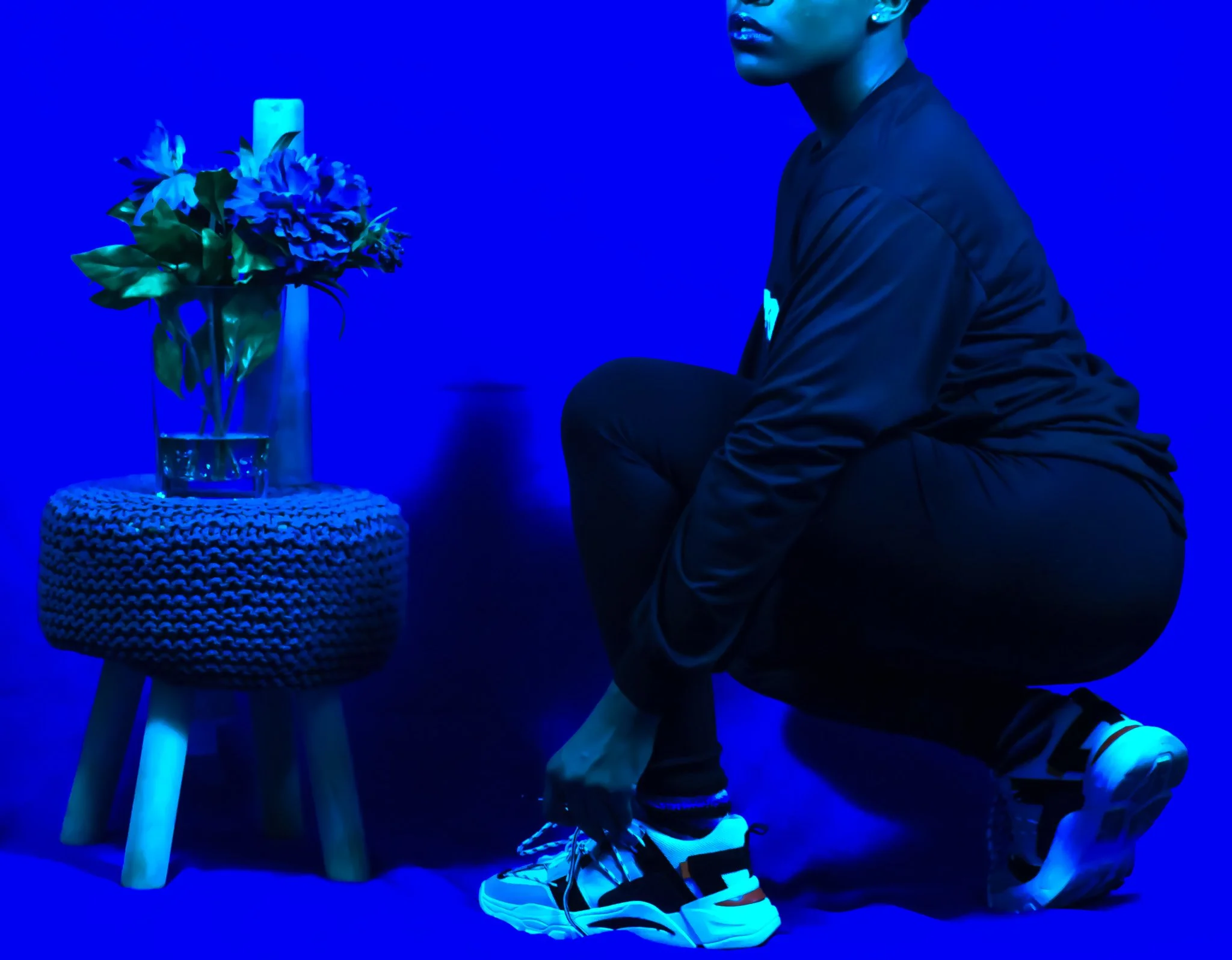 A person dressed in dark athletic clothing, squatting next to a small, round, textured stool with a glass vase of flowers on top, all illuminated by blue lighting.