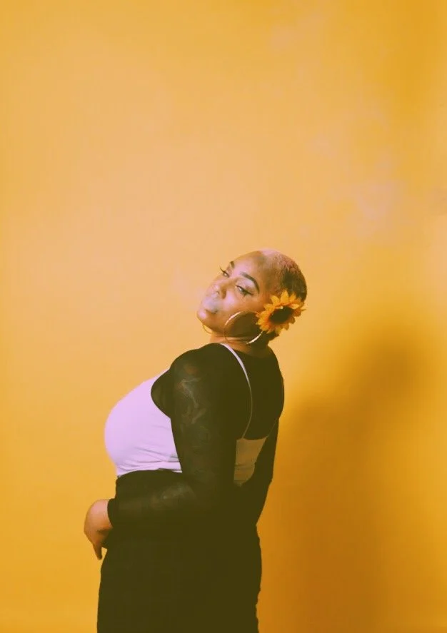 A woman with short hair and a sunflower behind her ear stands against a yellow background, wearing a black sheer long-sleeve top with a purple tank top underneath, and poses with her head tilted and eyes looking at the camera.