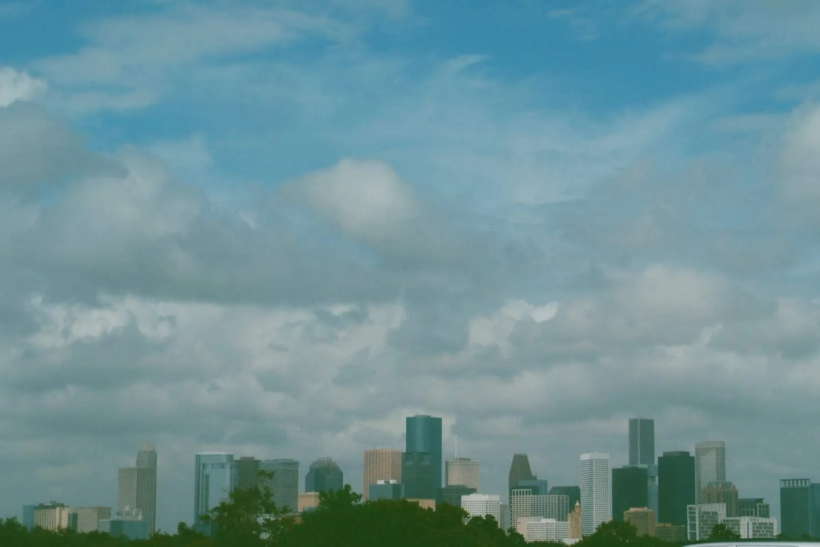 Skyscrapers in a city skyline under a partly cloudy sky with green trees in the foreground.