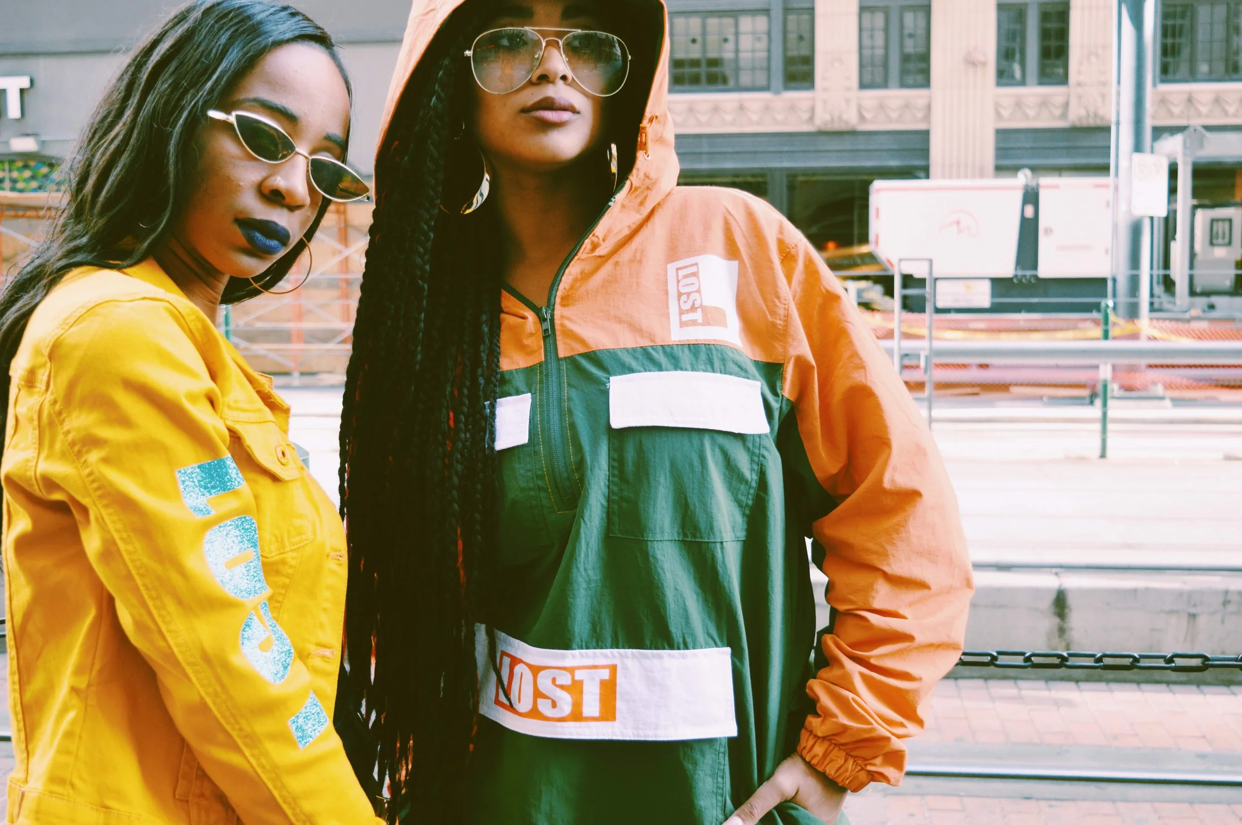 Two women standing outdoors in an urban setting, wearing colorful jackets and sunglasses, with a city street and buildings in the background.