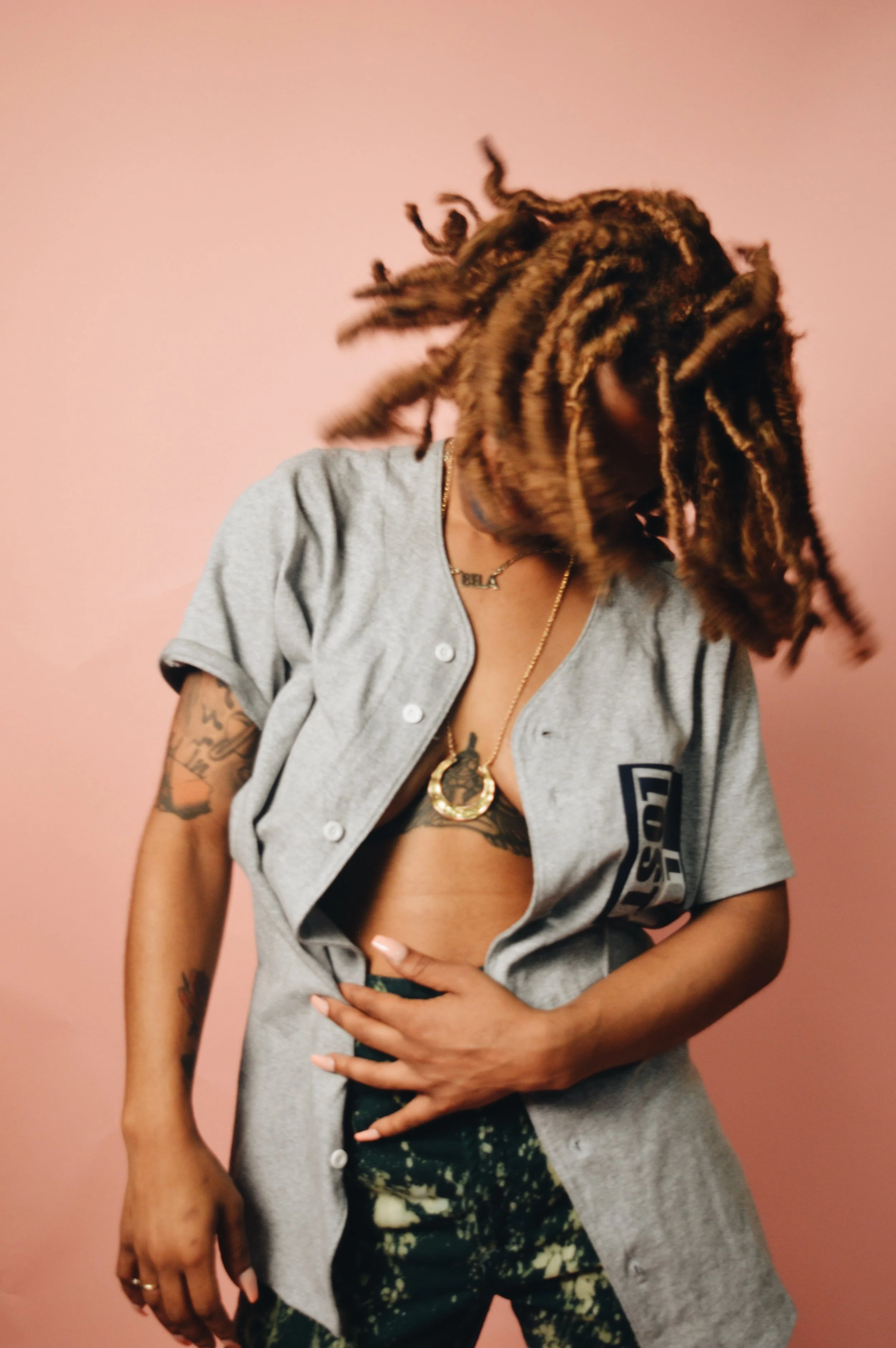 Person with dreadlocks wearing a gray sports jersey, exposing tattoos, gold jewelry, and black patterned pants, standing against a pink background.