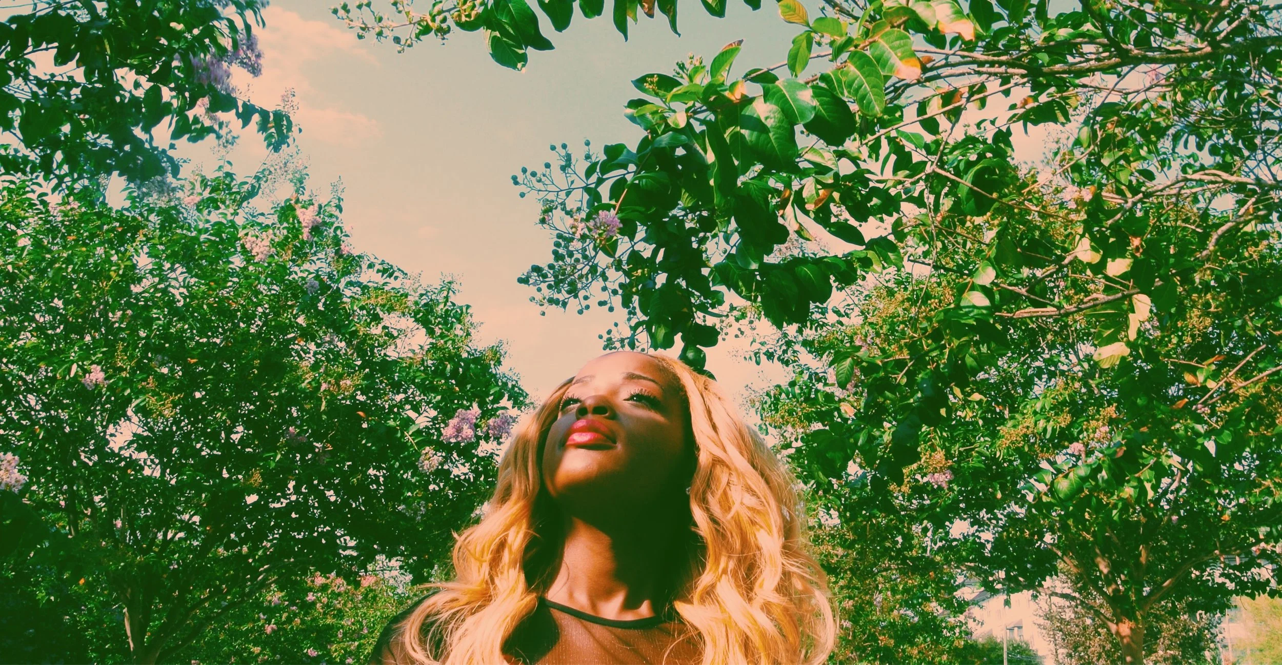 A woman with long blonde hair looking up at the sky surrounded by green trees and pink flowers.