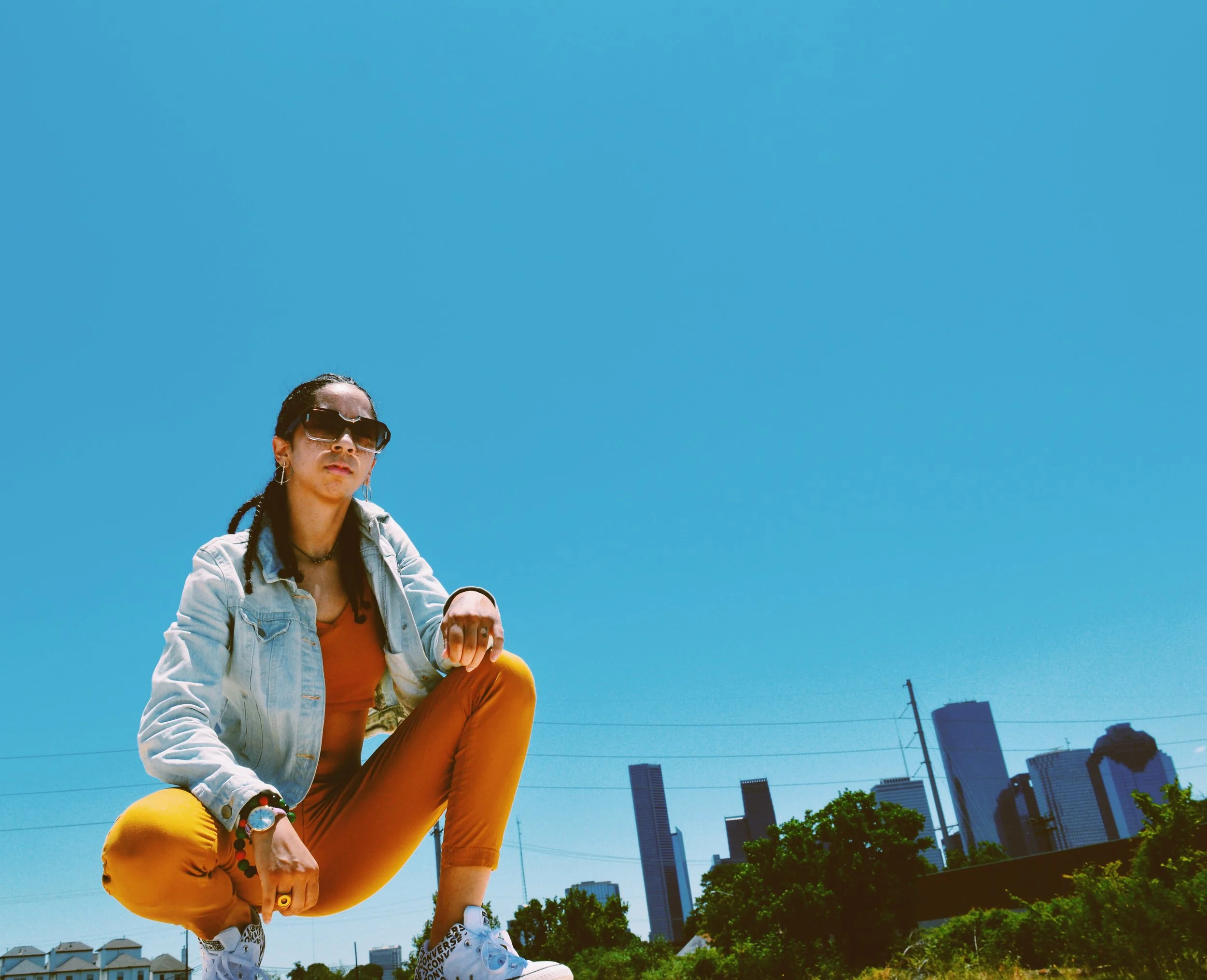 A person crouching outdoors against a clear blue sky with a city skyline with tall buildings in the background.