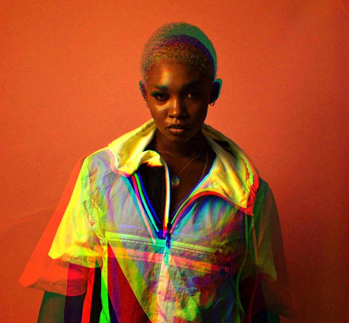 A woman with short, blonde hair wearing a black top and a shiny, multi-colored jacket, standing against a plain orange background.