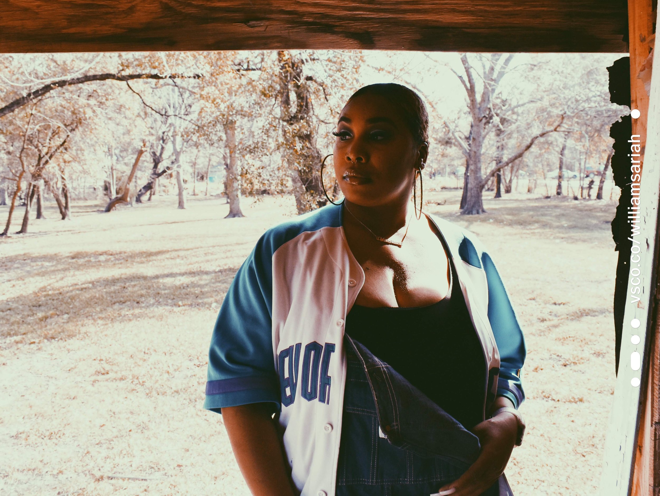 A woman standing outdoors on a sunny day with trees in the background. She is wearing a sports jacket, a black top, and hoop earrings.