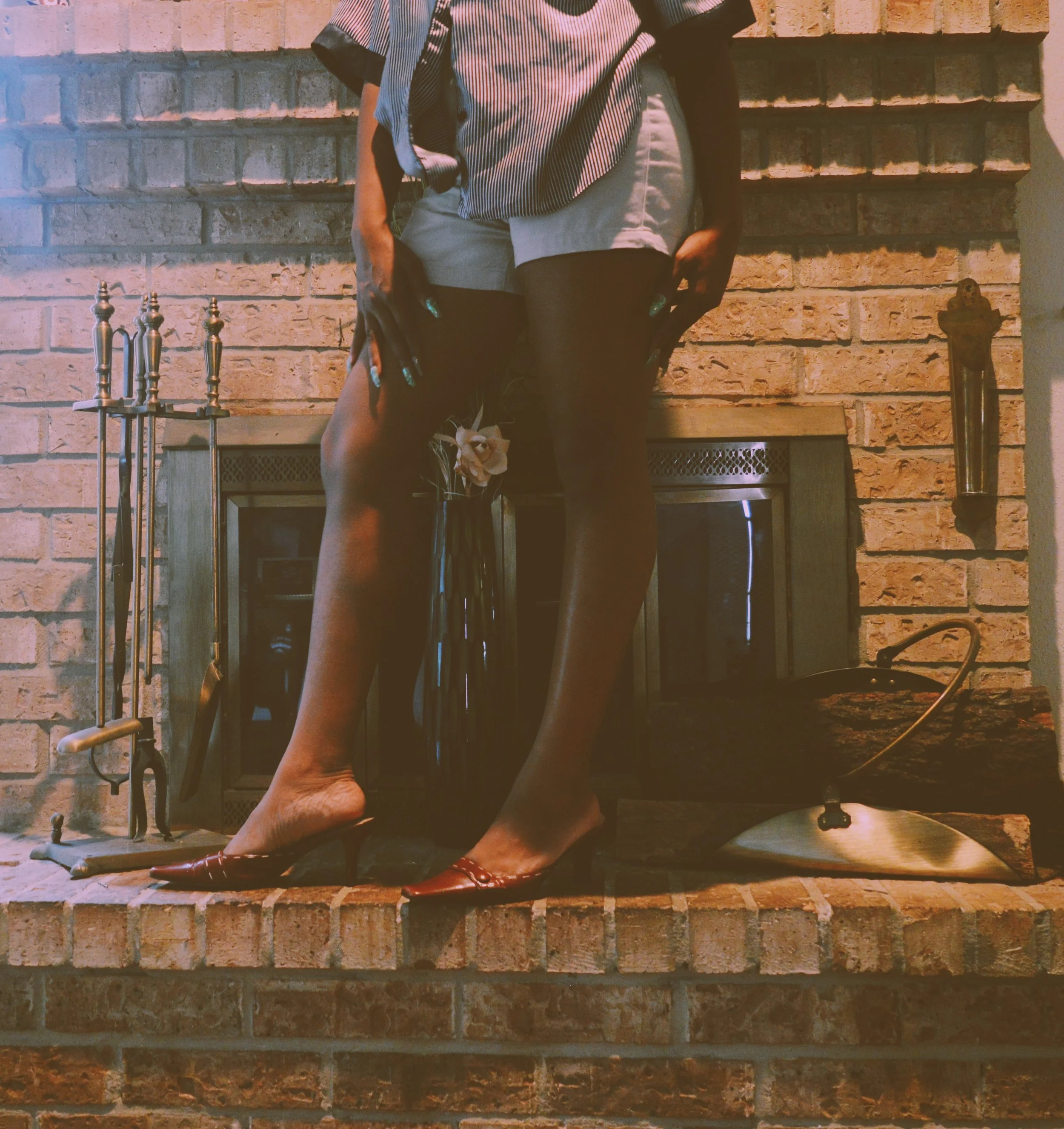 Close-up of a person standing on a brick fireplace mantle, wearing patterned heels, sheer black tights, and a striped blouse.