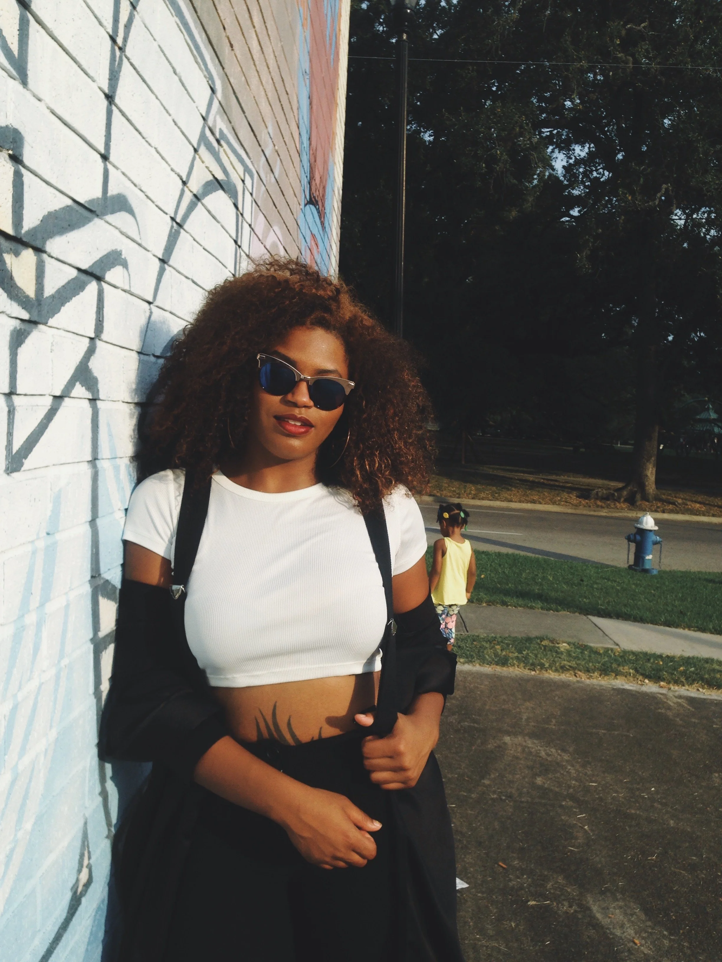 A woman with curly hair wearing sunglasses, a white crop top, and black jacket standing against a graffiti-covered wall during daytime. A child is seen in the background near a sidewalk and a fire hydrant.