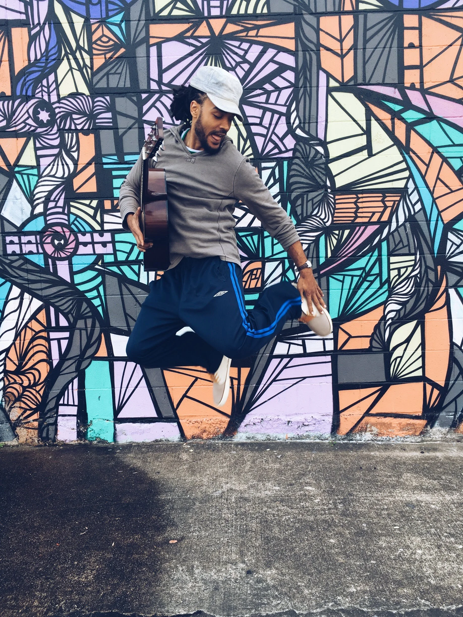 Man jumping in front of a colorful geometric graffiti mural while holding a guitar.