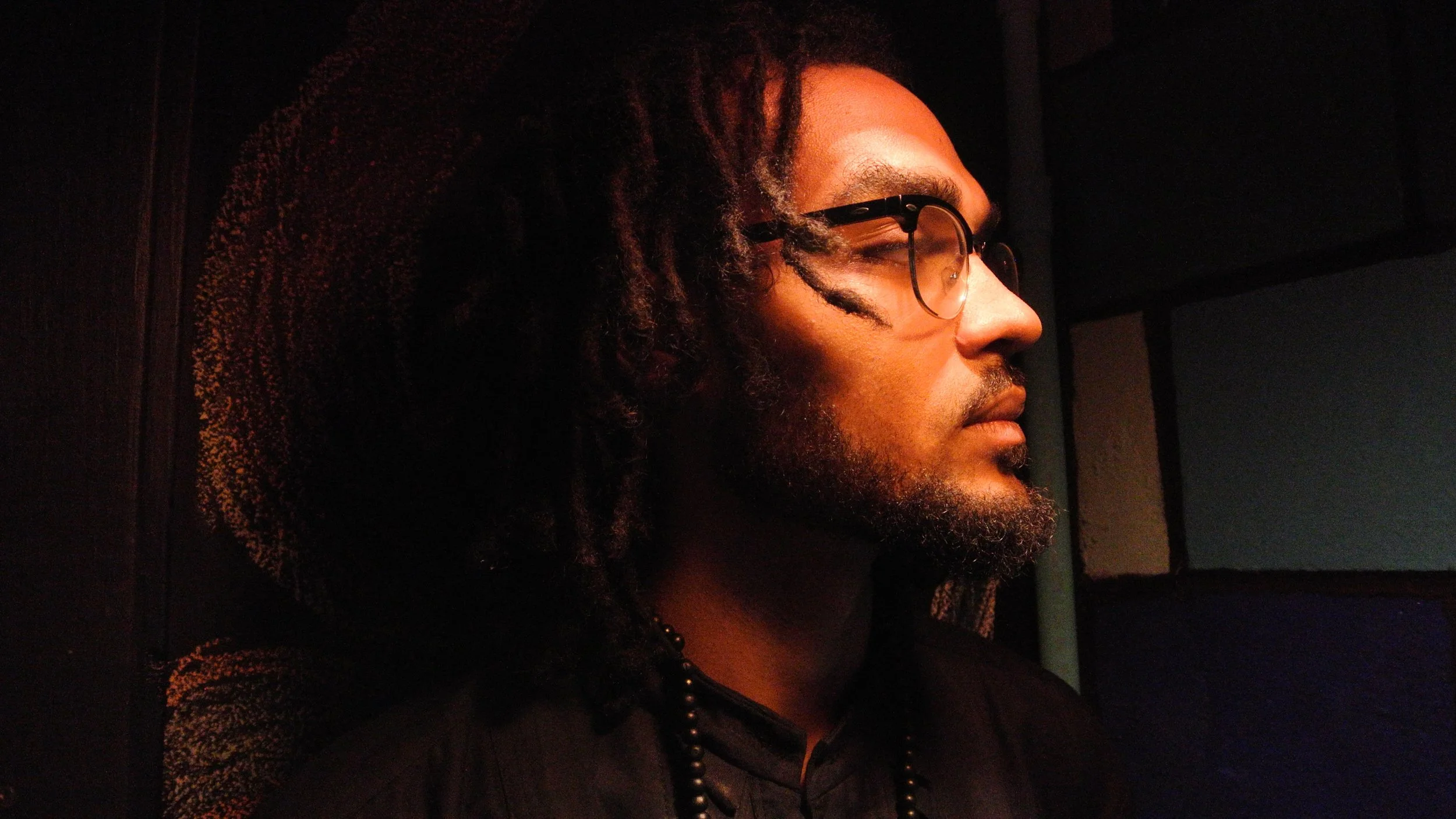 Profile of a man with dreadlocks and glasses, facing to the right, in dim lighting.