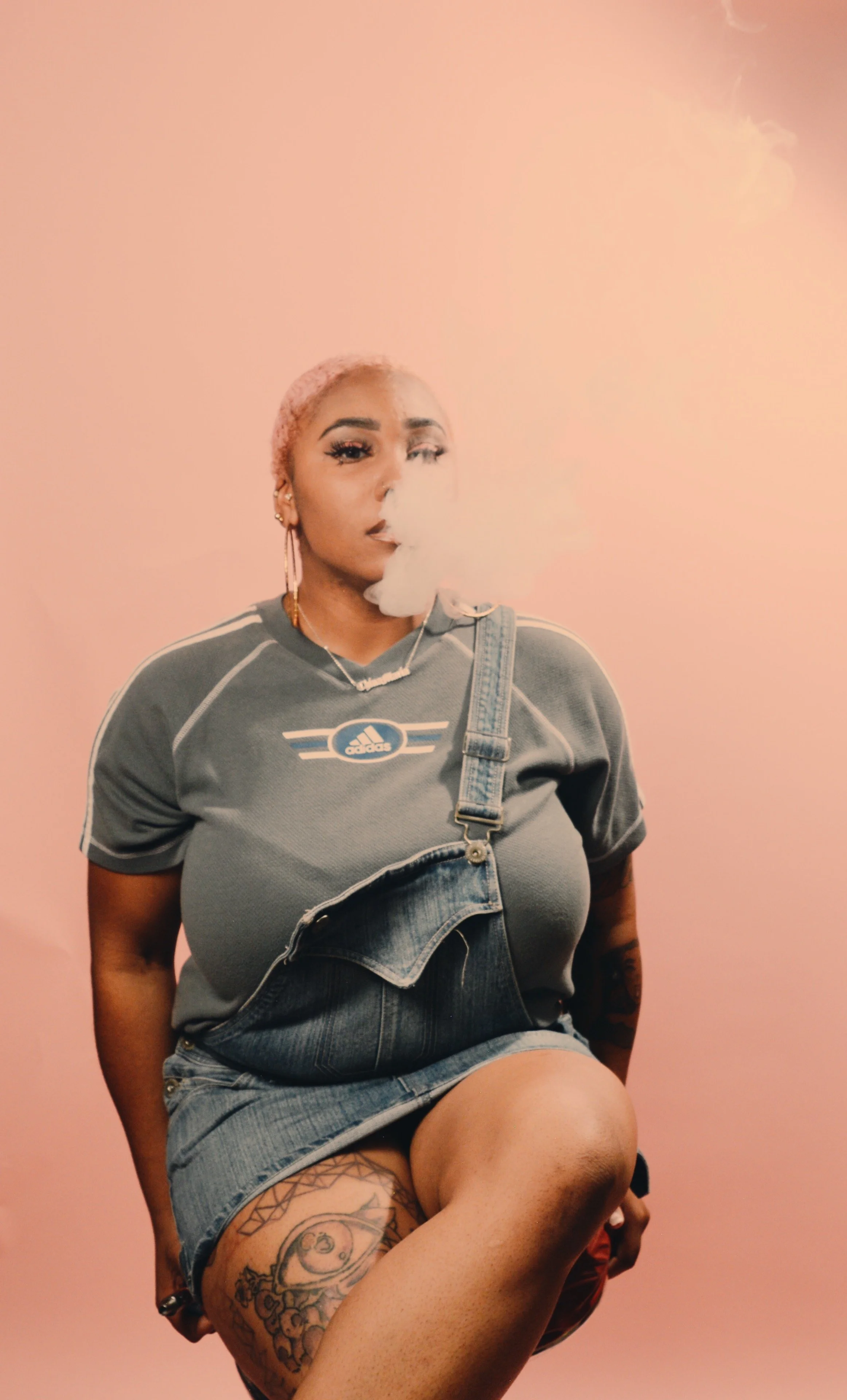 A woman with short curls and tattoos on her left thigh, sitting against a pink background, smoking vapor, wearing a gray Adidas t-shirt and denim overalls.