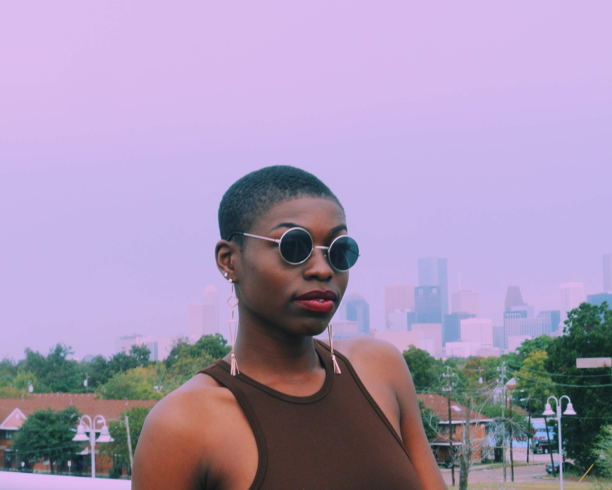 A woman with short hair, wearing sunglasses, earrings, and a brown tank top, stands outdoors with a city skyline in the background.