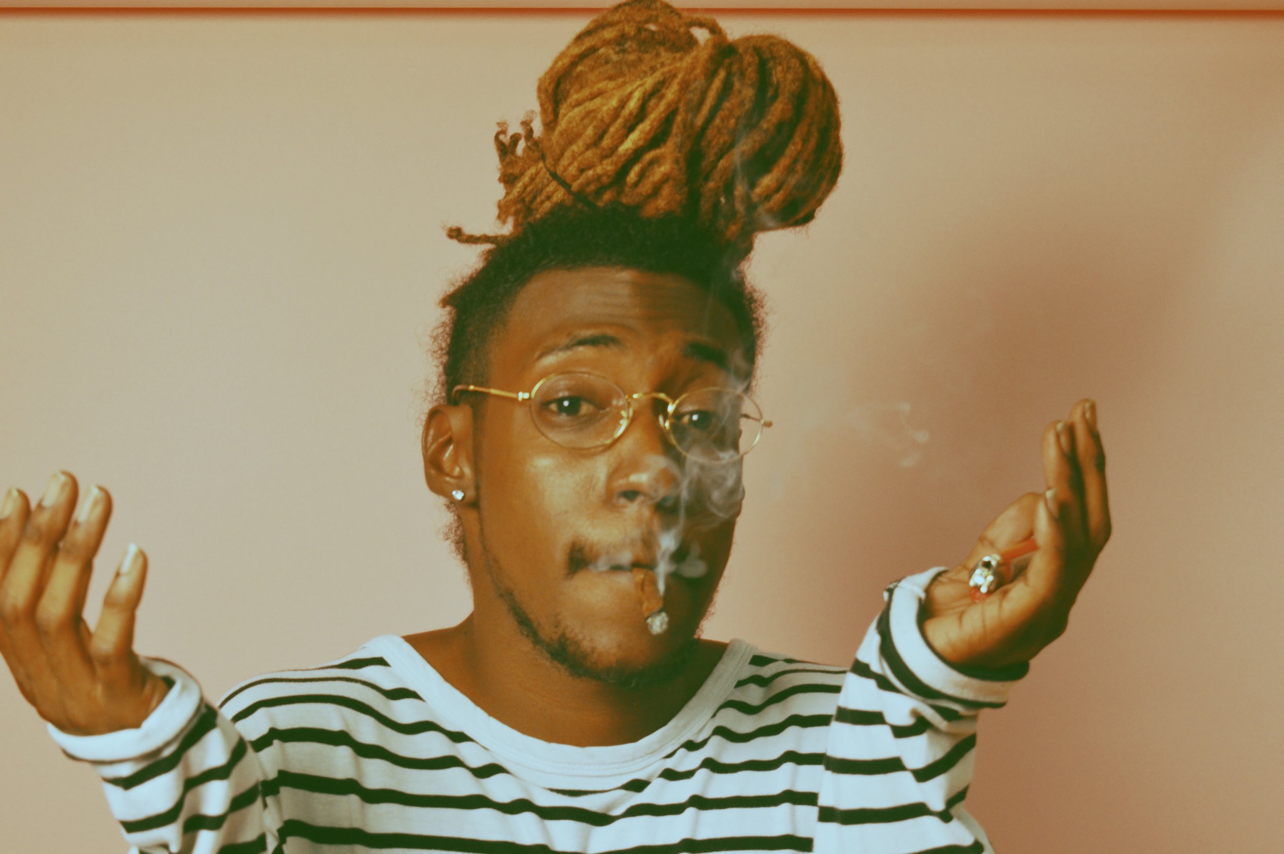 Person with dreadlocks styled on top, wearing glasses, smoking, and dressed in a striped shirt, standing against a plain wall.