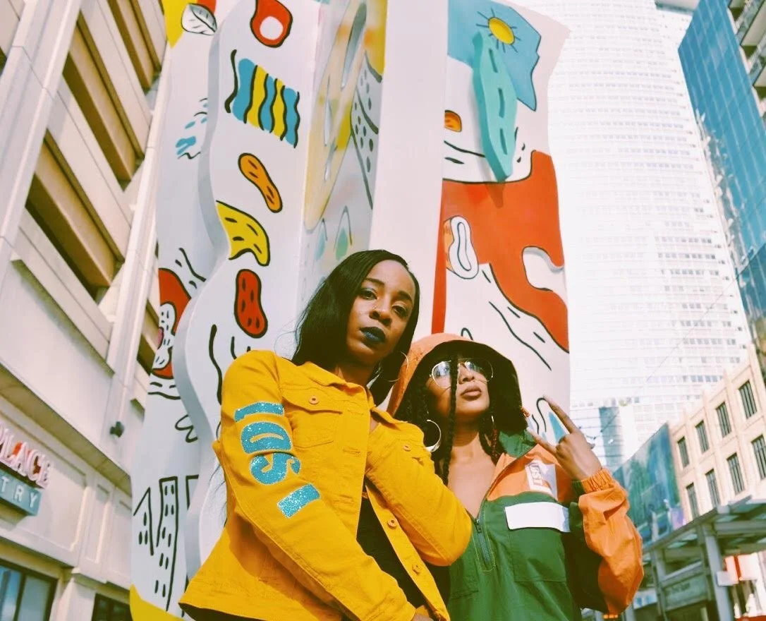 Two women standing in front of a mural with abstract and colorful shapes in an urban setting. One woman is wearing a yellow jacket and the other is wearing a multicolor jacket with glasses.