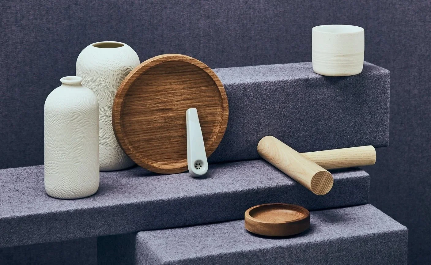 Collection of minimalist ceramic and wooden vases, bowls, and utensils arranged on gray textured display stands.