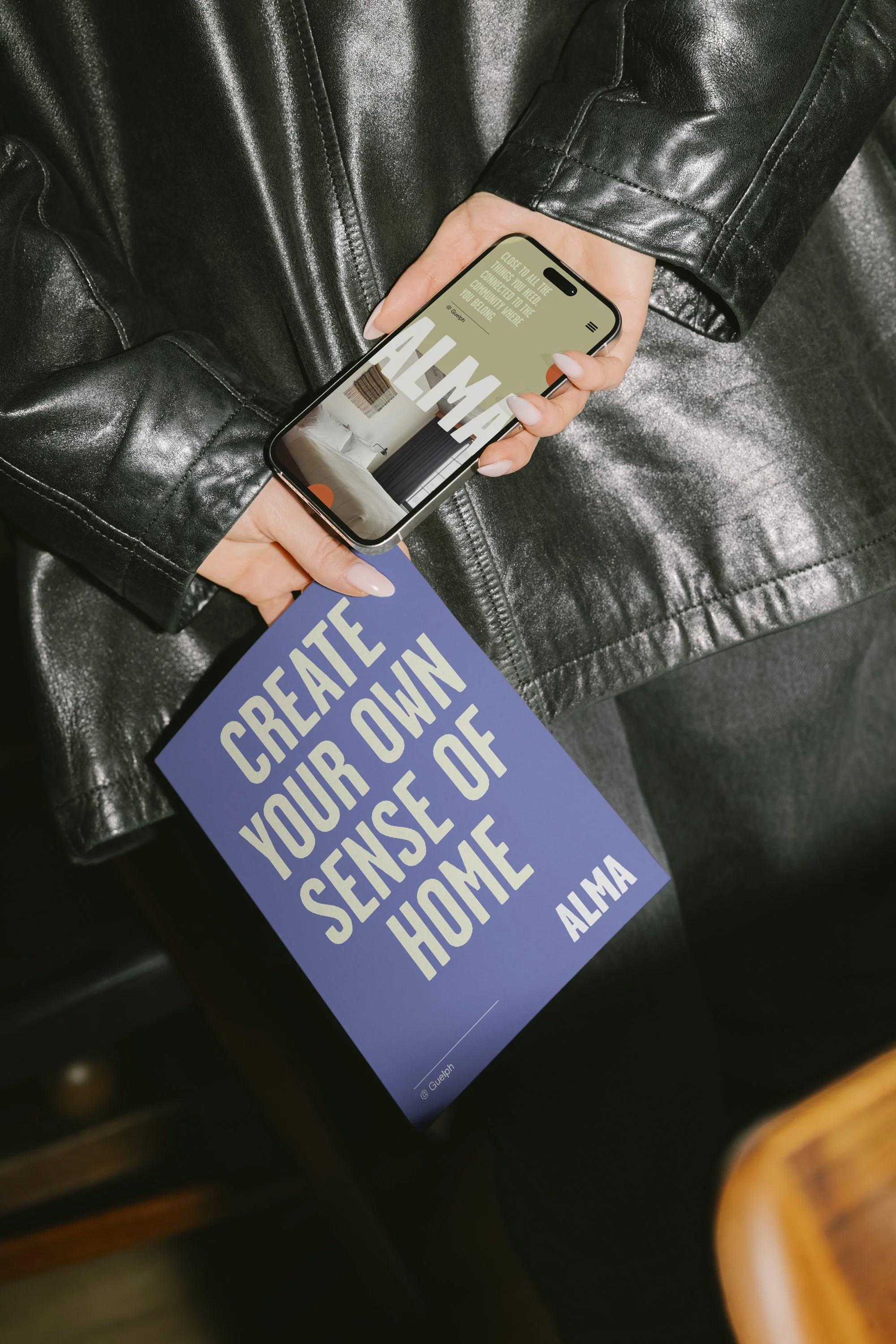 Person holding a purple sign with white text that says, "Create Your Own Sense Of Home - Alma," while wearing a black leather jacket and holding a smartphone displaying a website or app.