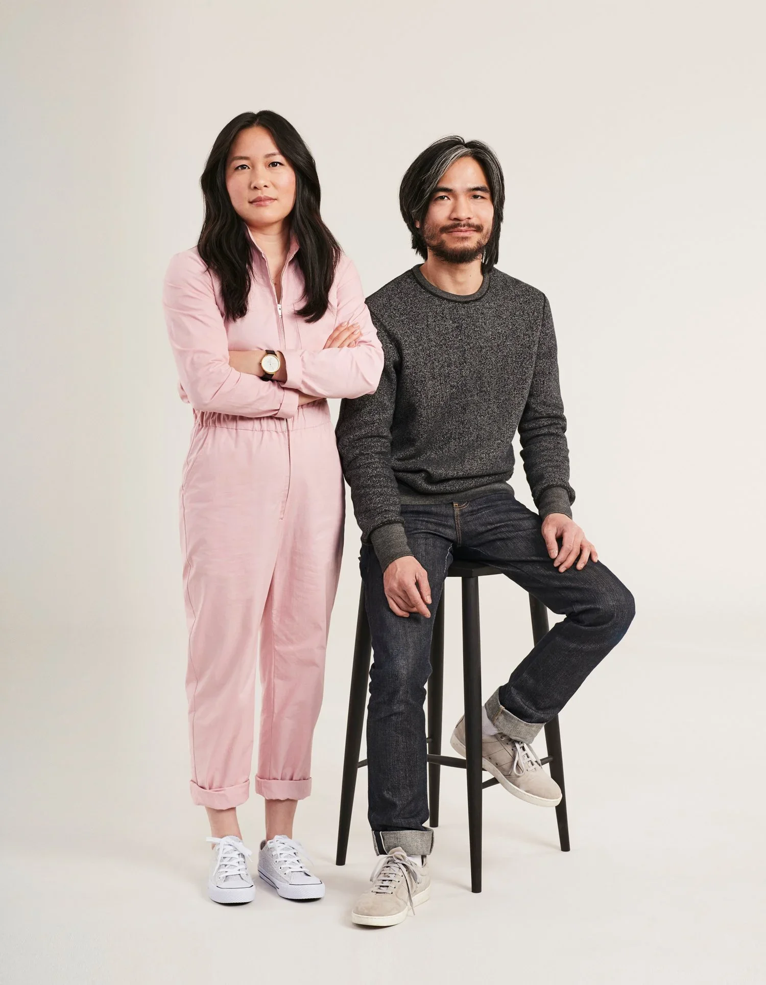 A woman in pink jumpsuit standing with arms crossed next to a man sitting on a stool, both facing forward against a plain background.