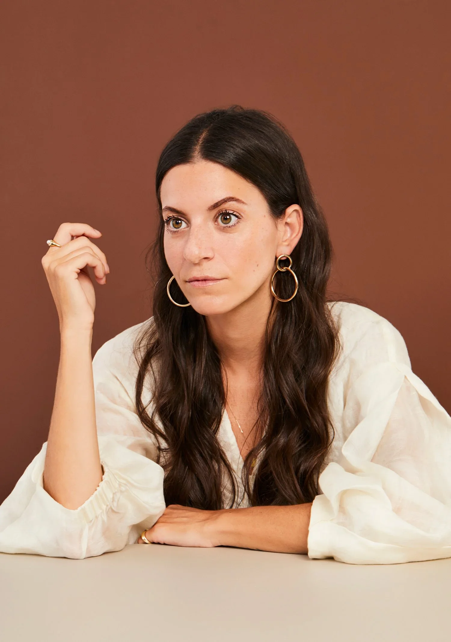 A woman with long dark hair, wearing gold hoop earrings and a cream-colored blouse, sitting at a table against a brown background.