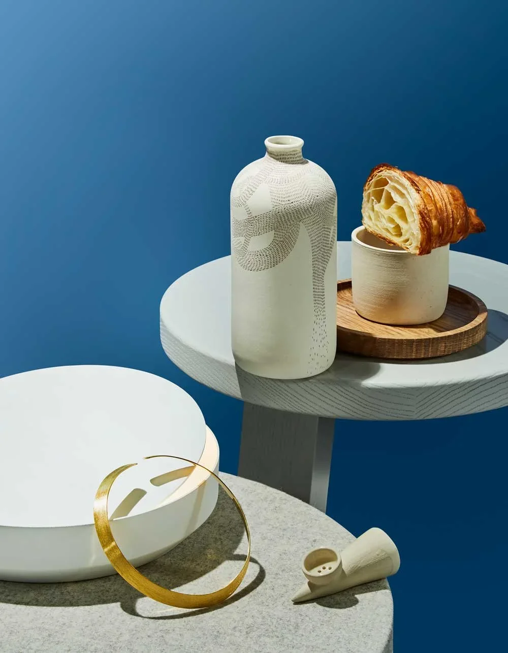 A still life scene featuring a white ceramic bowl with a gold metal ring on top, a white ceramic pitcher with a textured design, a small white cup, a wooden tray, and a croissant in a white cup on a wooden saucer, against a blue background.