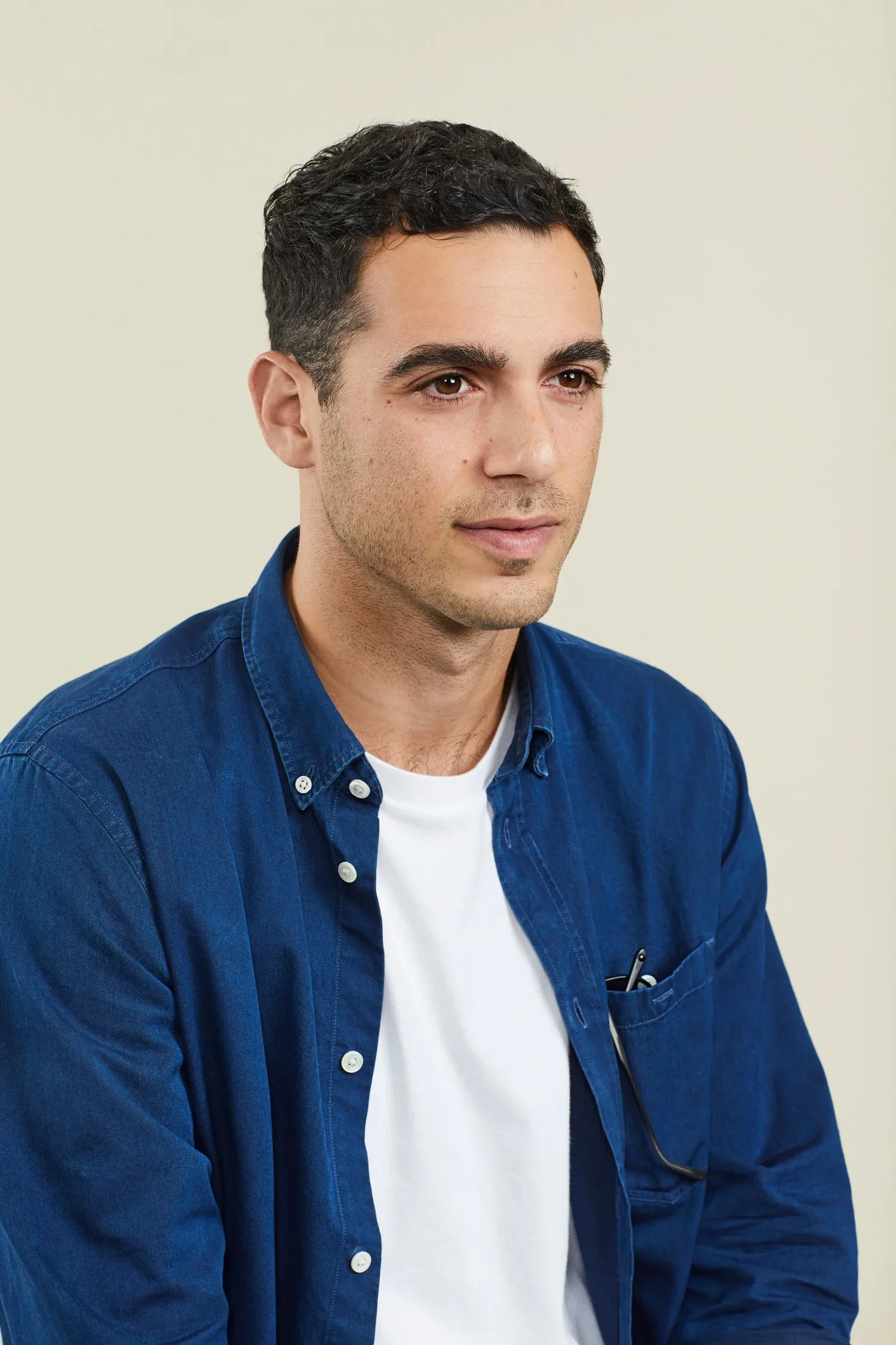 A young man with dark hair, wearing a blue button-up shirt and white t-shirt, sitting against a plain light background.