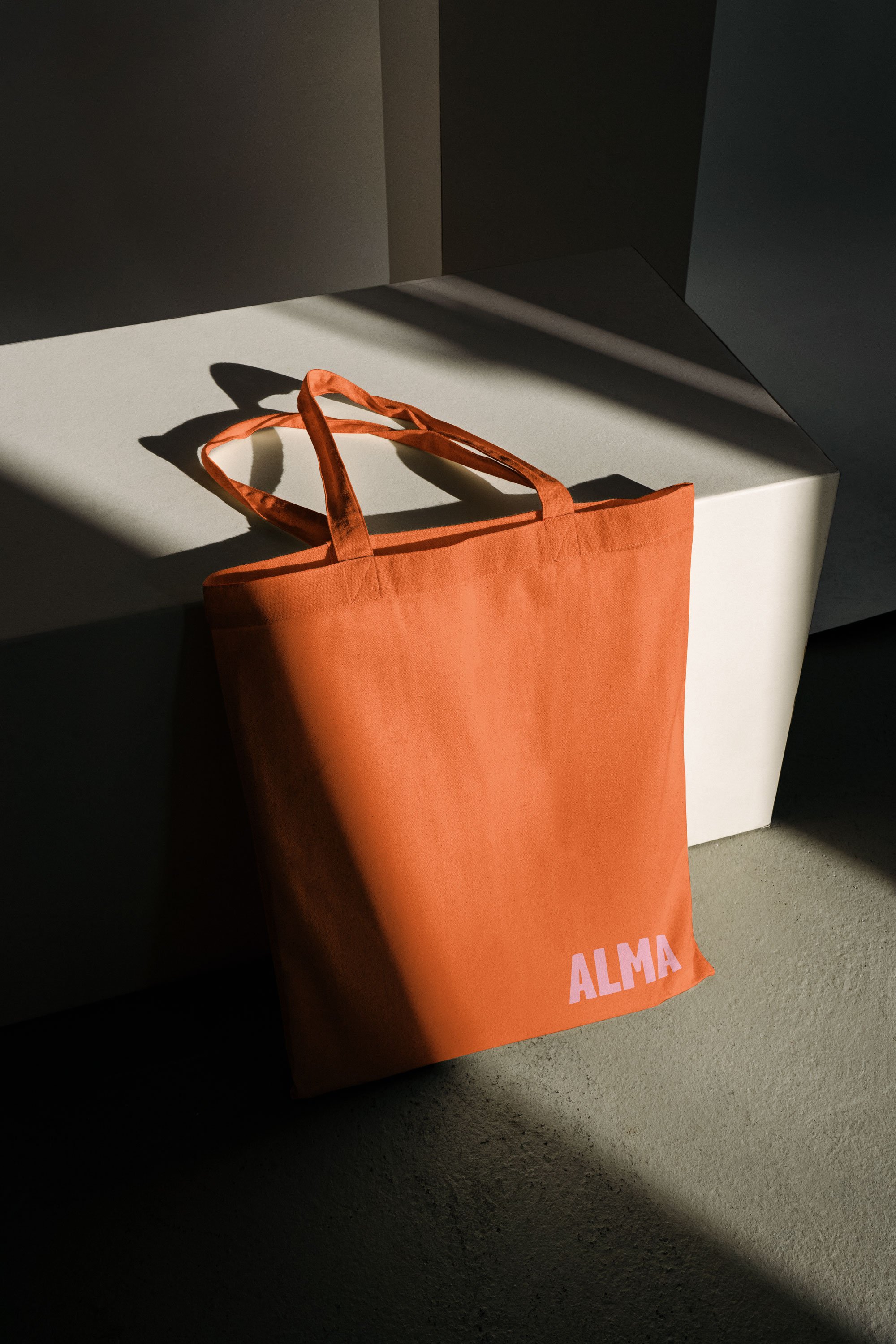 Orange tote bag with the word 'ALMA' printed in white, resting on a white surface with a shadow cast across it.