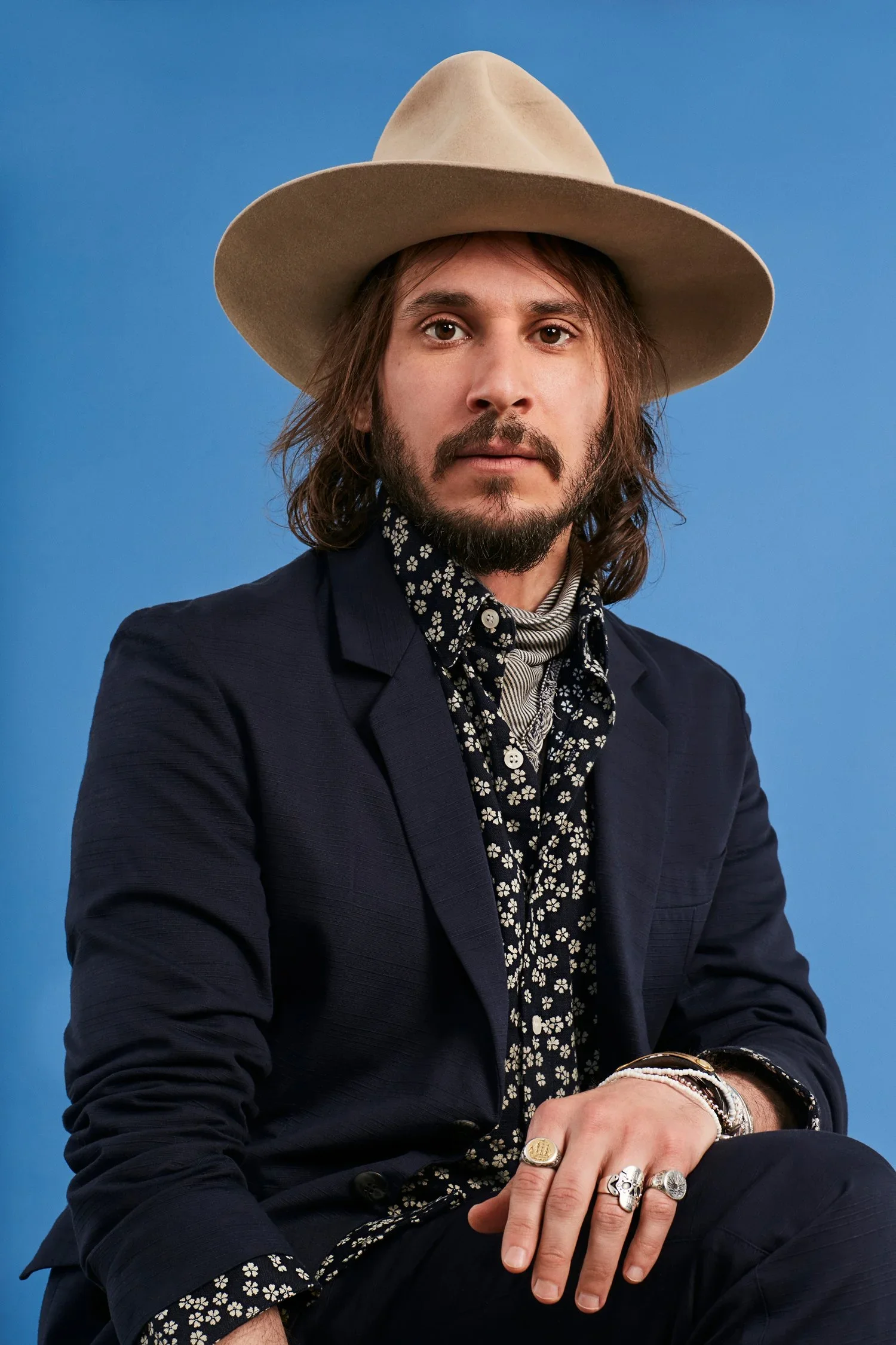 Man with long brown hair, beard, wearing a beige wide-brimmed hat, navy blazer, black shirt with white floral pattern, sitting against a blue background.