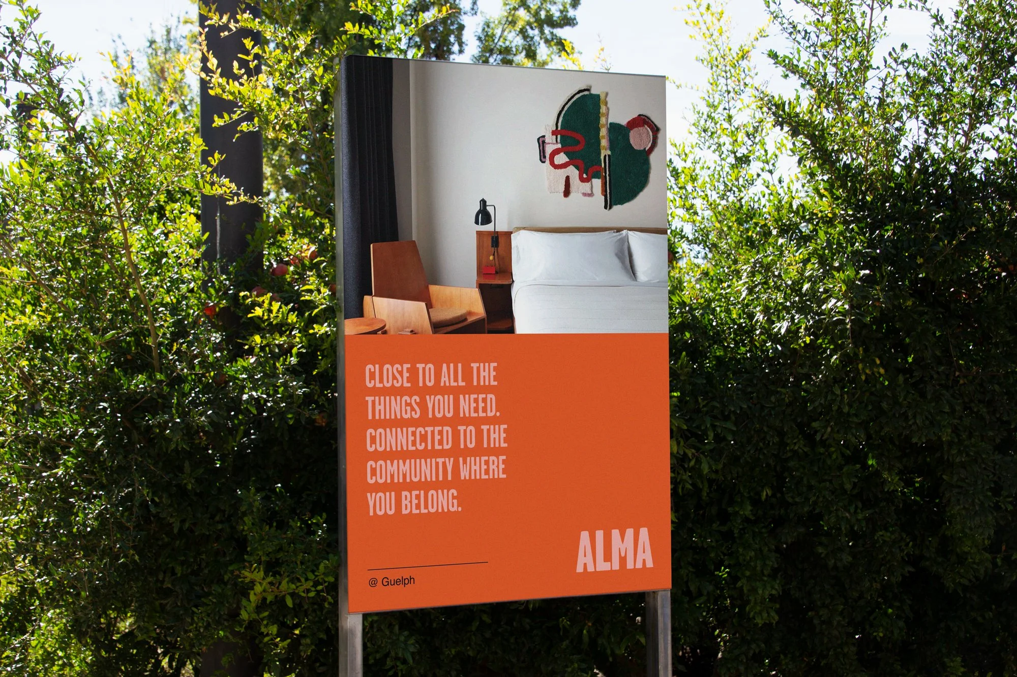 Outdoor sign with an interior room photograph at the top, showing a bed, a nightstand with a lamp, a wooden chair, and a wall art piece. The bottom section of the sign is orange with white text that reads: "Close to all the things you need. Connected to the community where you belong. ALMA" and includes the handle "@ Guelph."