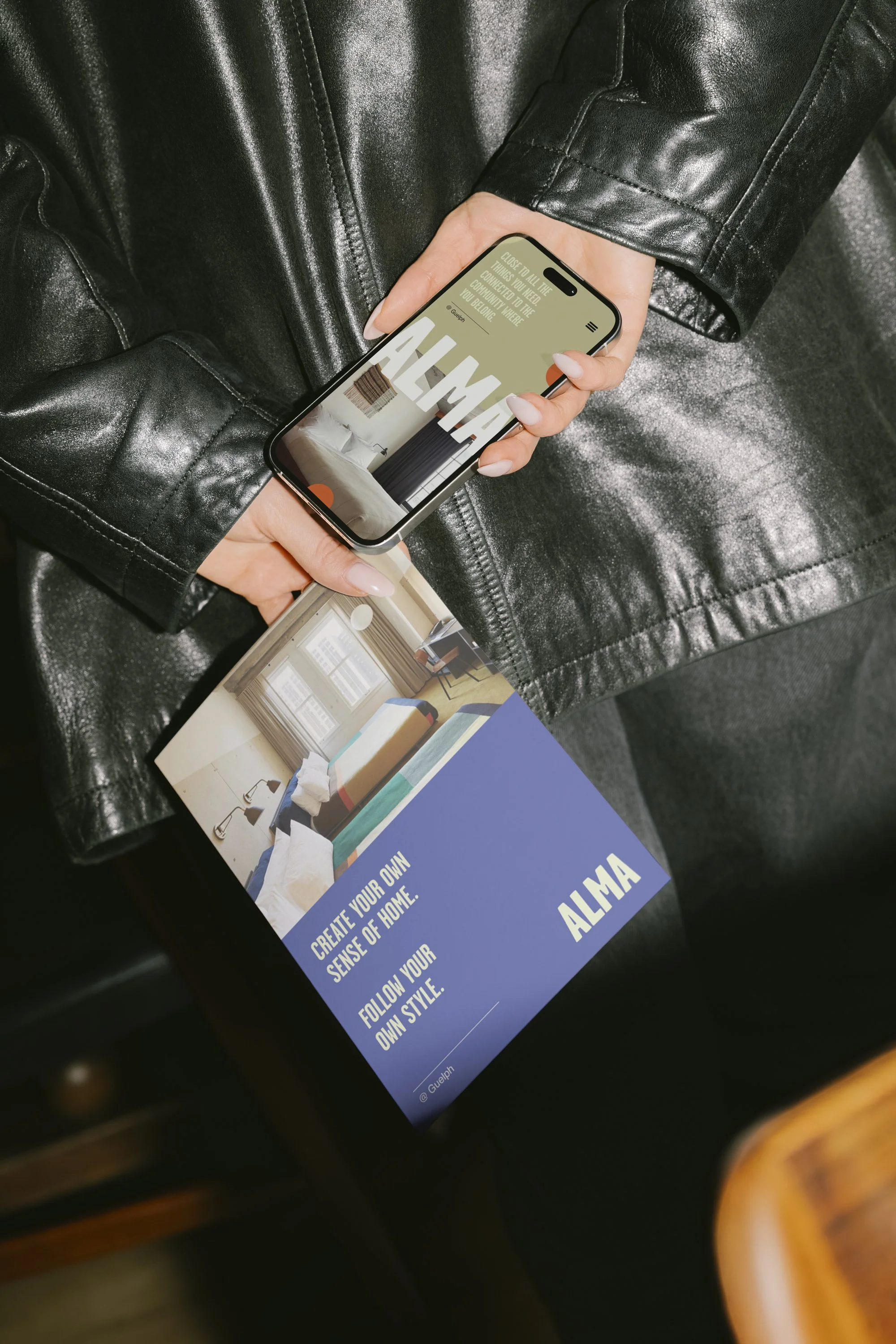 A person wearing a black leather jacket holding a smartphone and a brochure. The brochure shows a bedroom interior and has the text 'Create your own sense of home. Follow your own style. ALMA'.