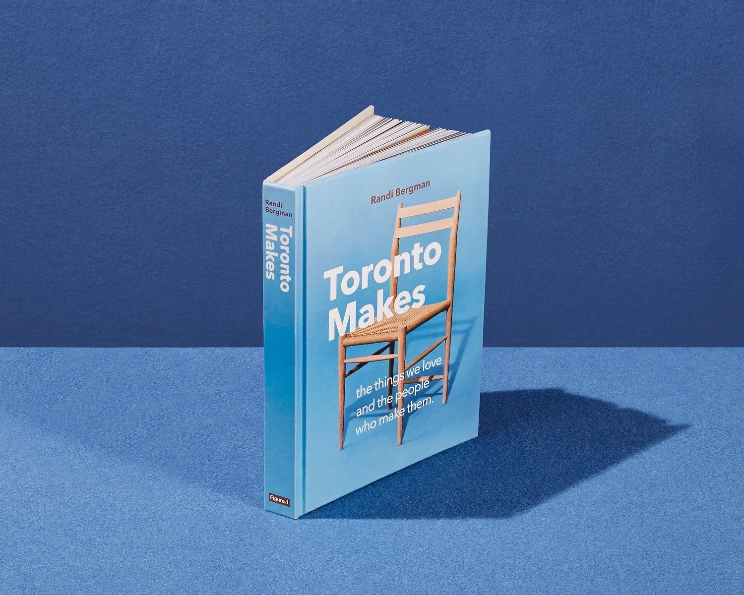 A book titled "Toronto Makes" standing upright on a blue surface, with a blue background. The cover features an image of a wooden chair. The author's name, Randi Bergman, is at the top.