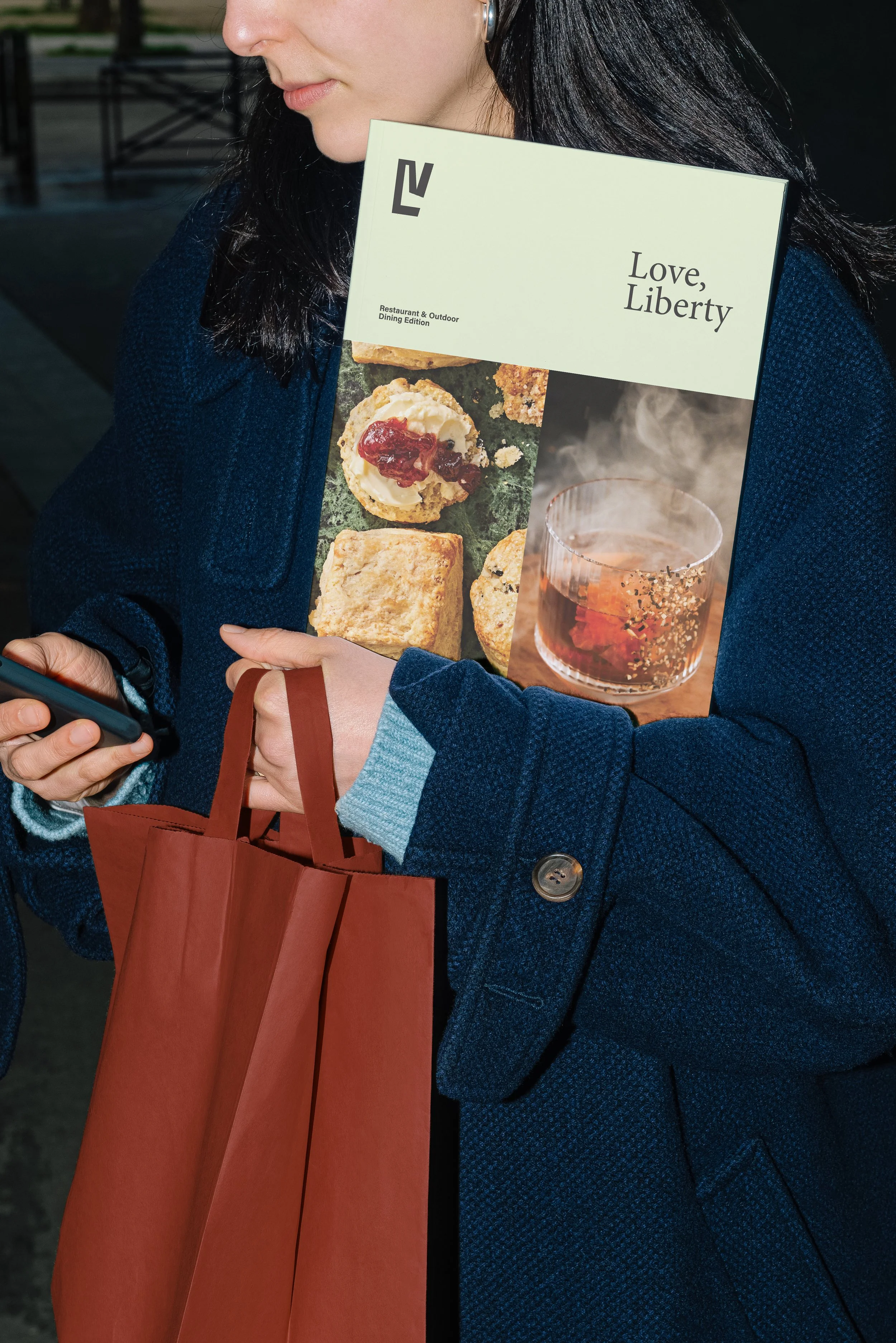 A woman wearing a blue coat holding a red shopping bag and a restaurant menu while looking at her phone.