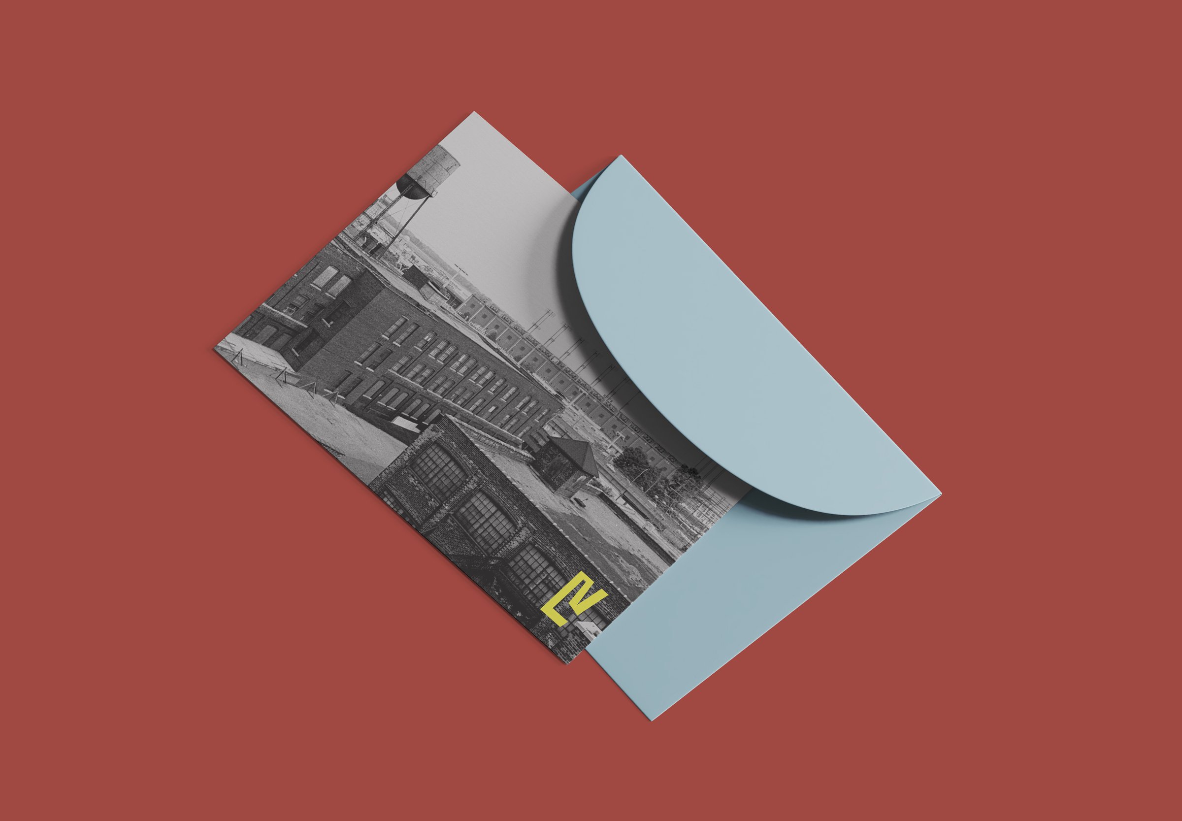 A folded postcard showing a black and white photo of an brick industrial building and a water tower, with a yellow 'E' logo in the bottom right corner. The postcard is partially pulled out from a light blue envelope on a red background.