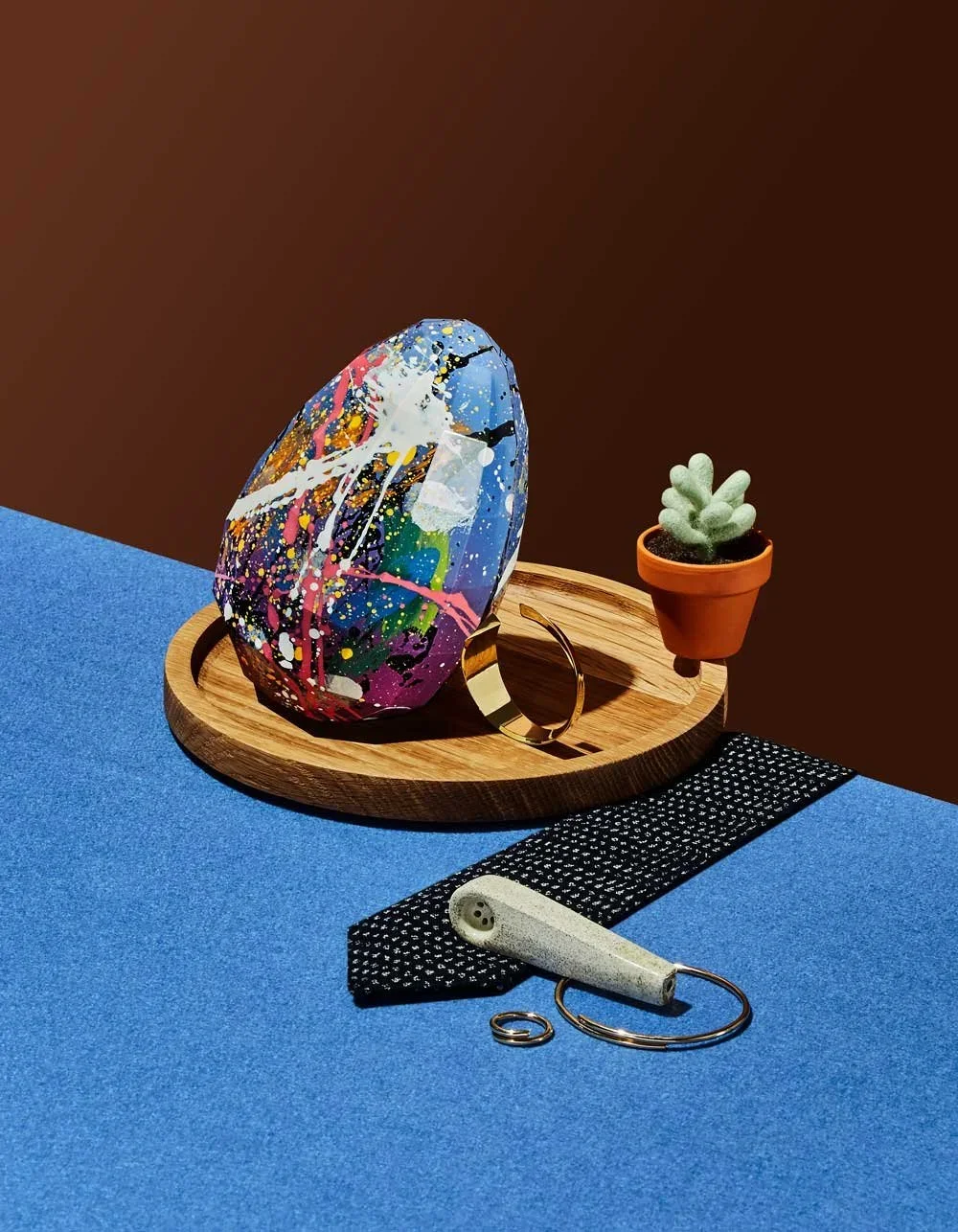 Colorful abstract glass paperweight on a wooden tray, with a small potted succulent and a reflective bracelet or ring, set on a blue surface with a brown background.