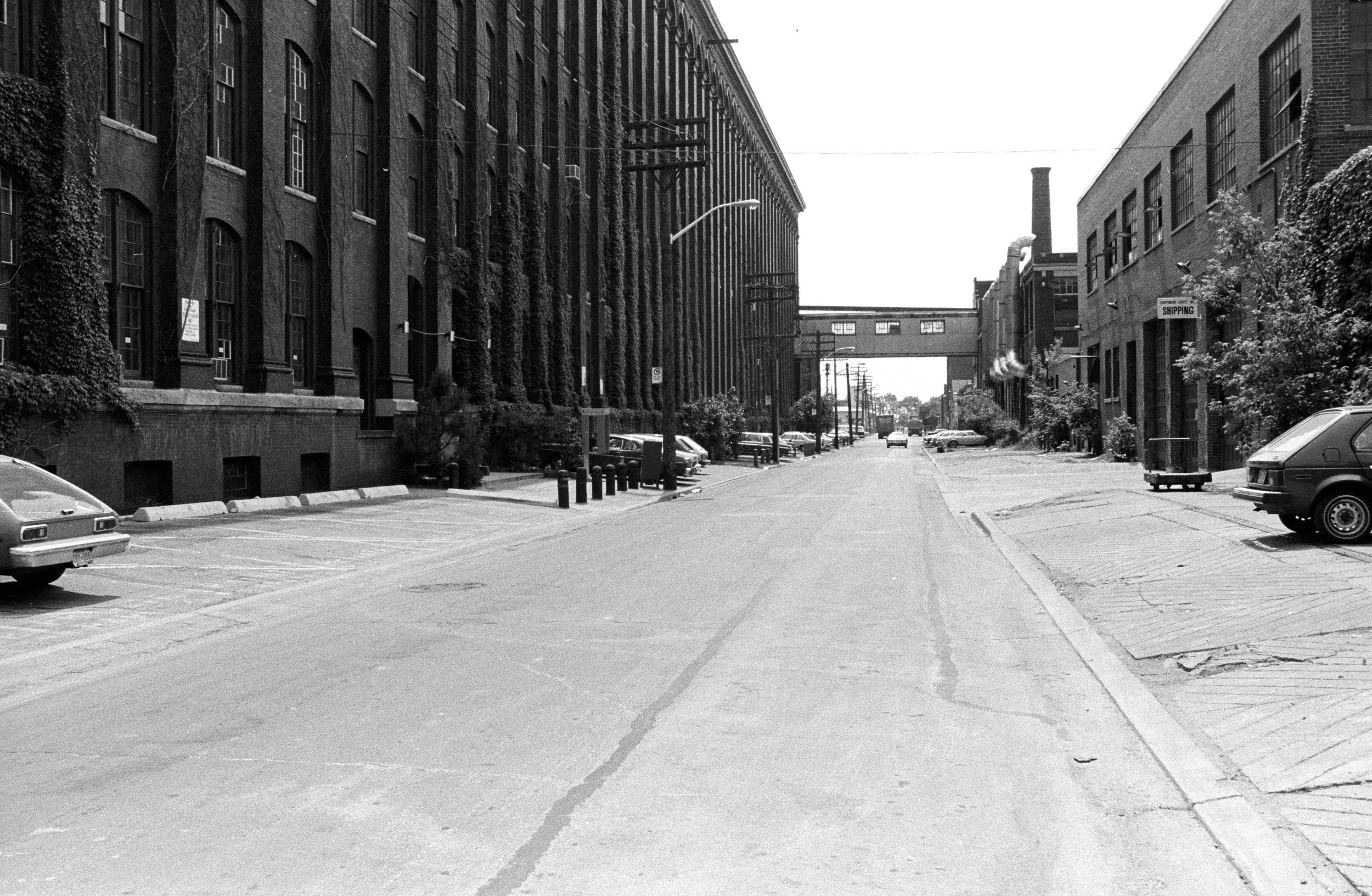 A black and white photo of an empty urban street with parked cars, industrial buildings, trees, and a pedestrian bridge in the distance.