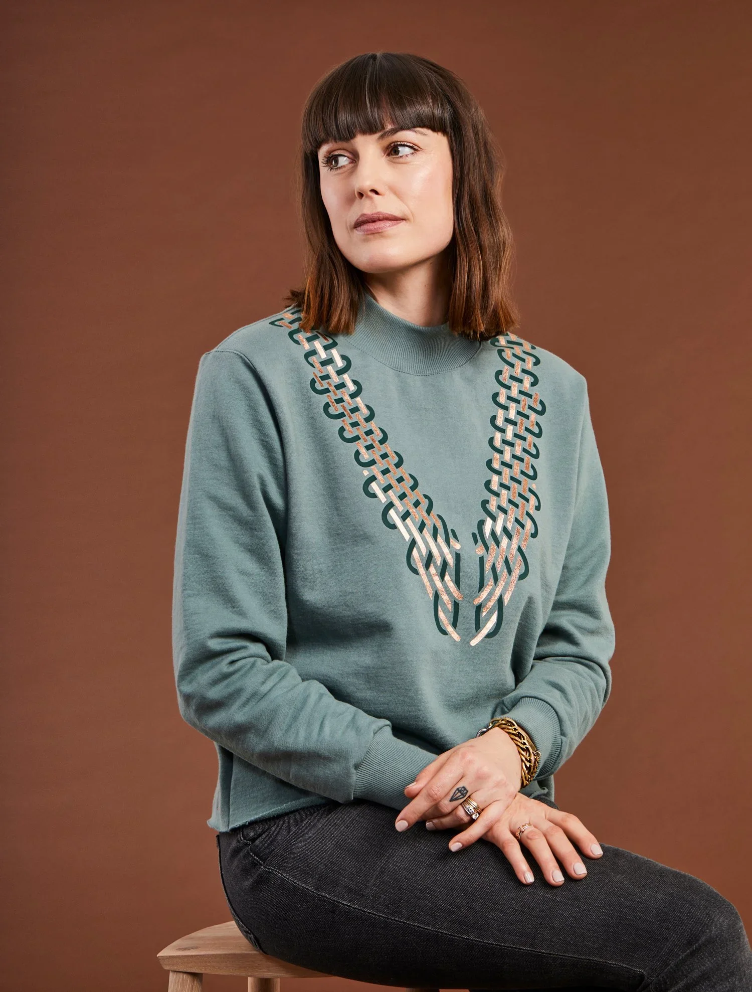 A woman with shoulder-length brown hair and bangs sits on a wooden stool against a plain background. She wears a grey sweatshirt with a chain pattern on the front and has several rings and bracelets on her hands and wrists.