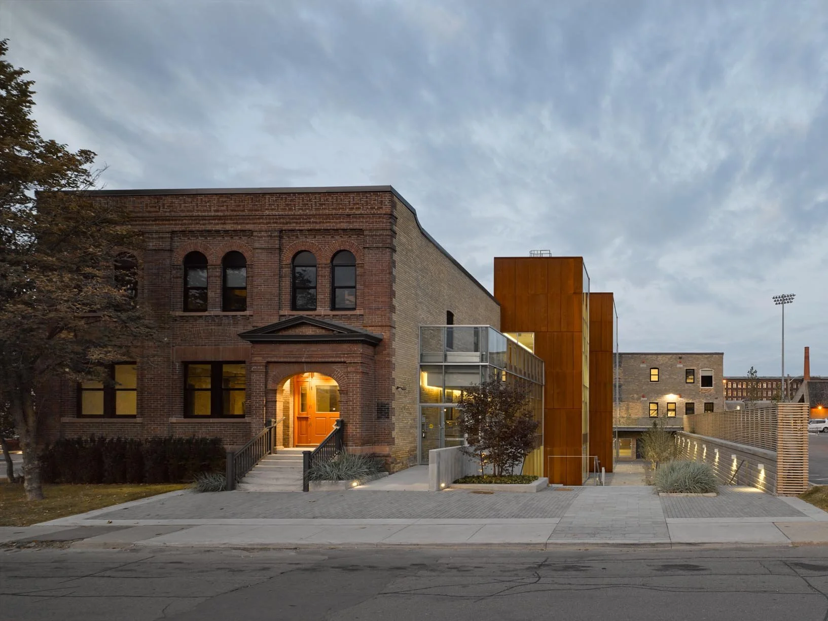 A modern building with a brick facade and a glass extension, featuring a rust-colored metal structure, at dusk with interior lighting visible and a partly cloudy sky overhead.