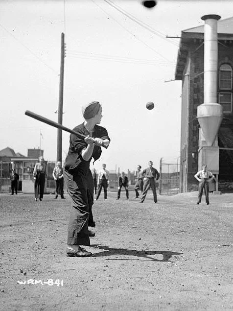 A young person wearing a turban and clothes is playing baseball, swinging a bat at a ball. Several other people are watching or participating in the background near an industrial building and street poles.