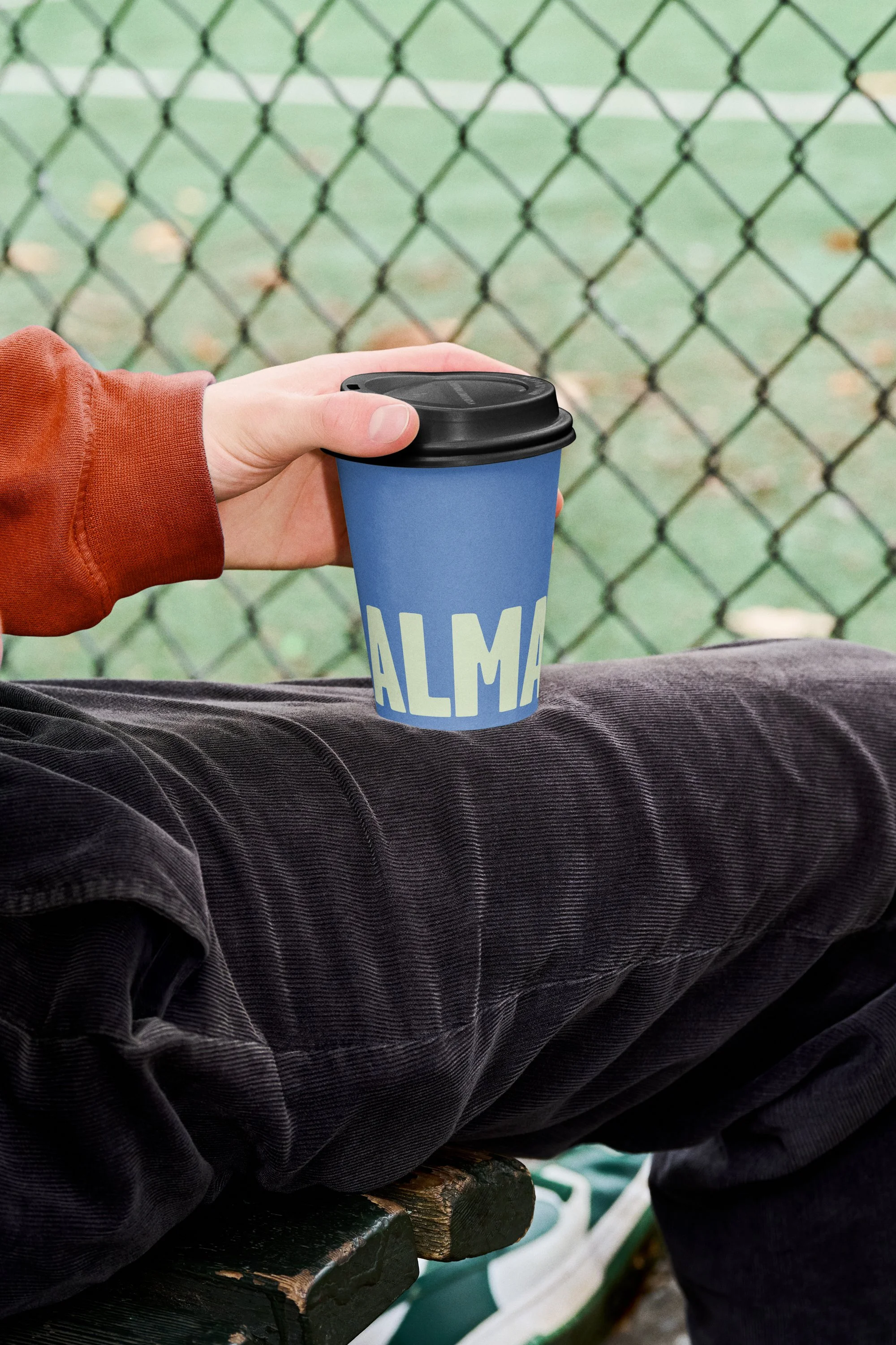 A person sitting on a park bench holding a blue paper coffee cup with a black plastic lid, near a chain-link fence, with a green sports field in the background.