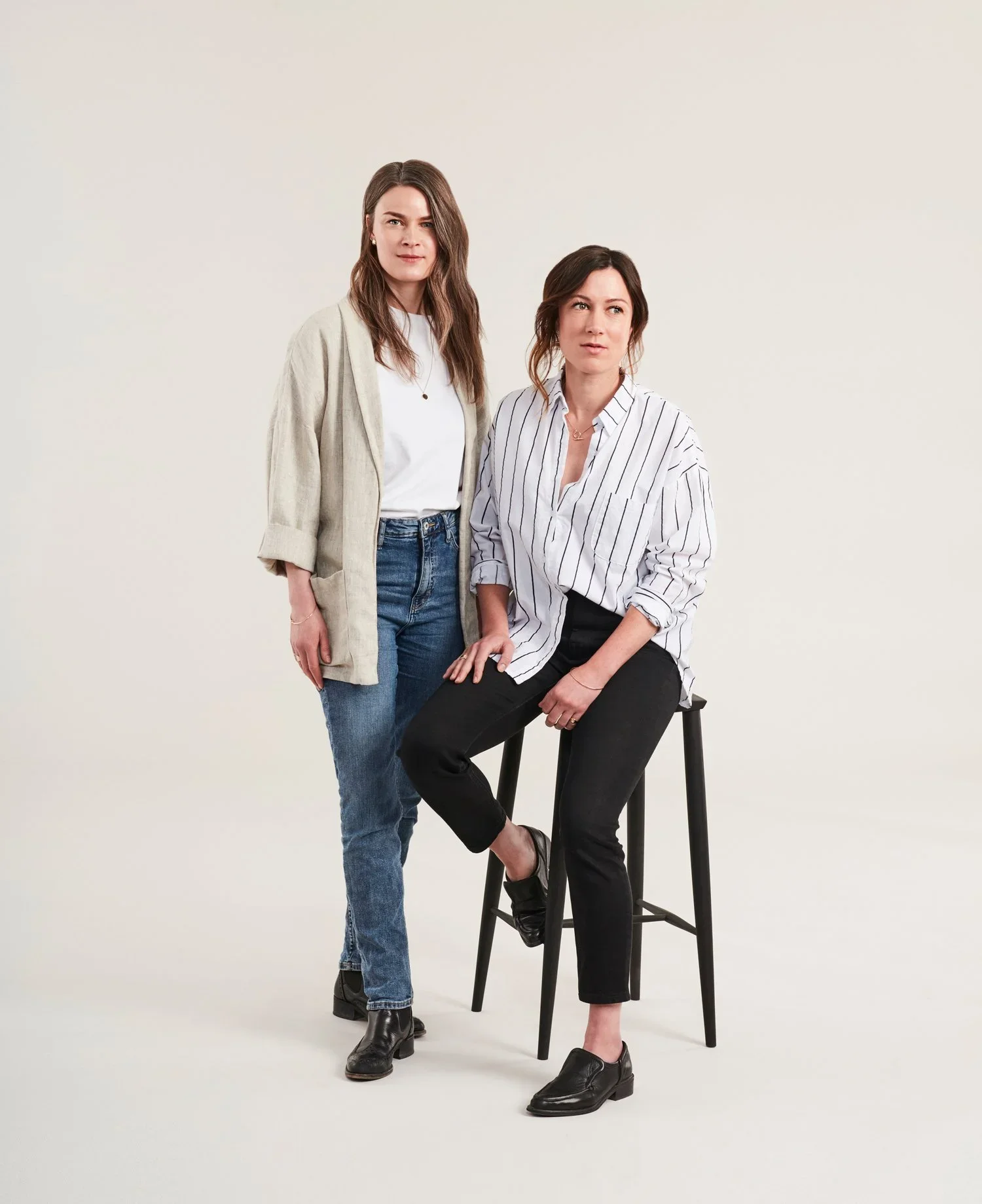 Two women with brown hair, one standing and one seated on a tall stool, against a plain light background.
