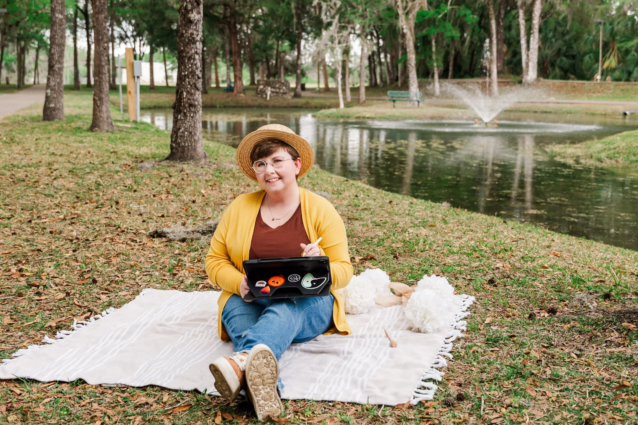 A woman sitting on a white blanket by a pond in a park, holding a tablet with Halloween stickers, smiling, wearing a straw hat, brown shirt, yellow cardigan, glasses, and blue jeans. There are paper lanterns and a stick nearby, with trees and a fountain in the background.