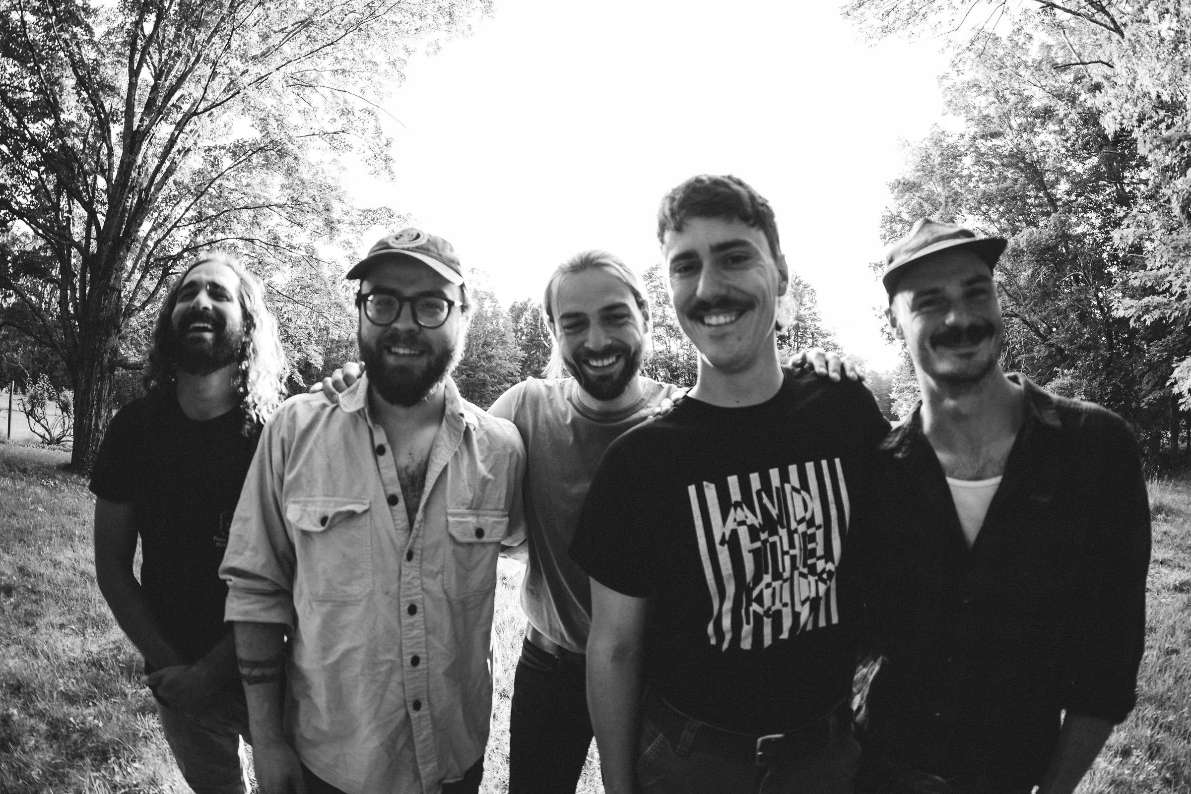 Six smiling men standing outdoors in a park during daytime, surrounded by trees.