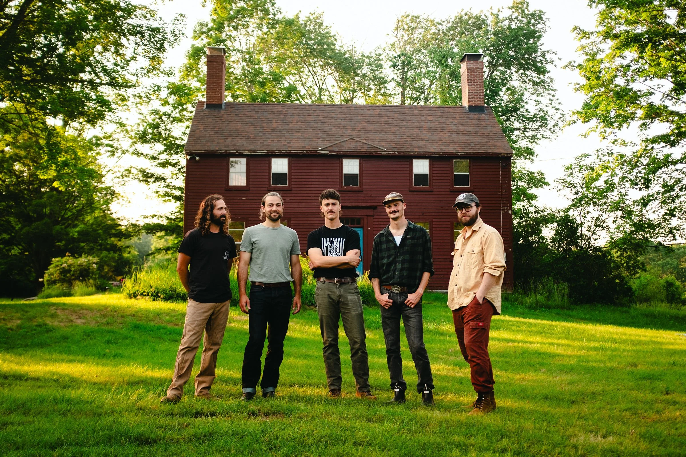 Group of five men standing on a lawn in front of a red wooden house surrounded by trees.