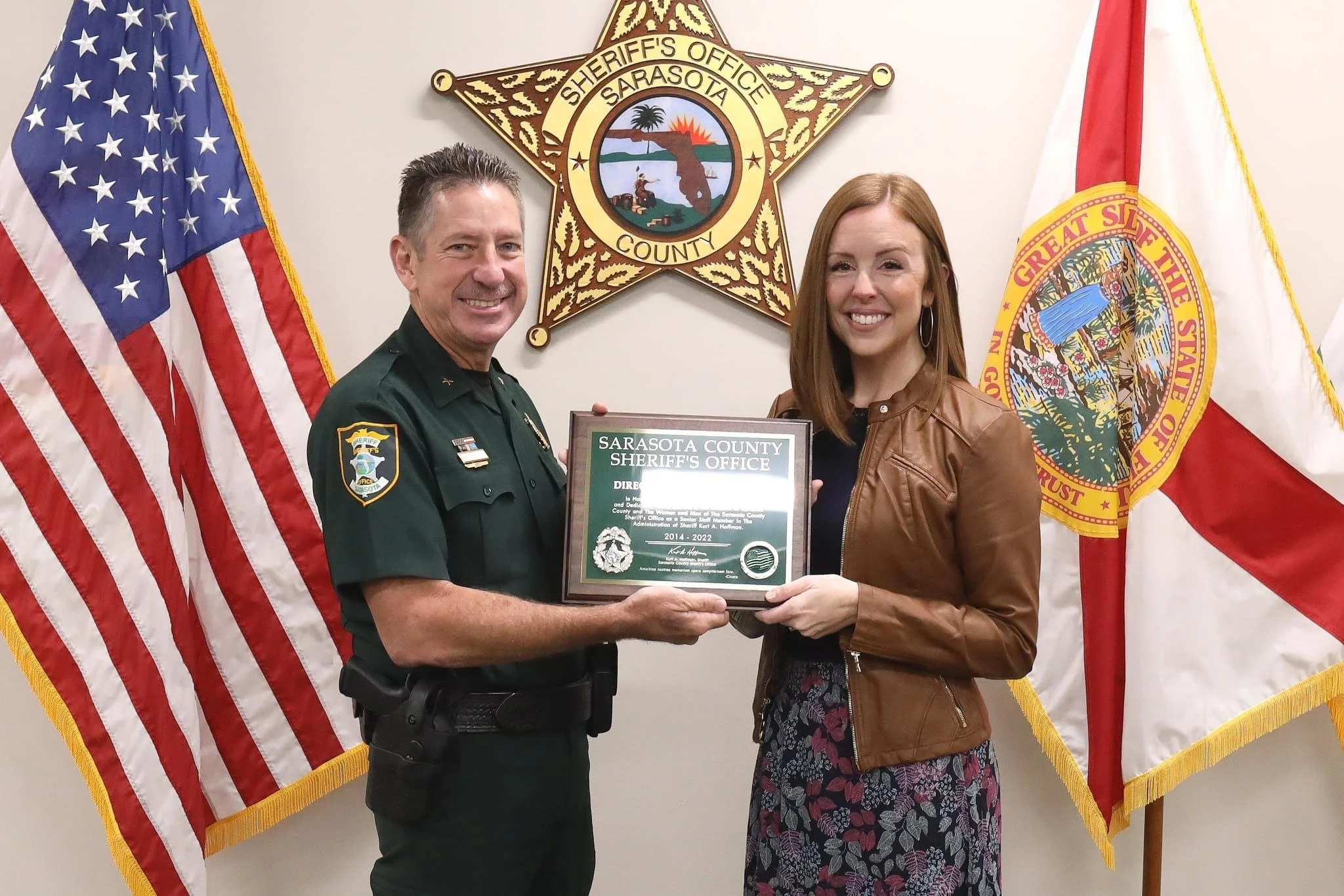 A man in a sheriff's uniform and a woman in a brown jacket holding a framed award at the Sarasota County Sheriff's Office. The background features the American flag, the Sarasota County Sheriff badge, and the Florida state flag.