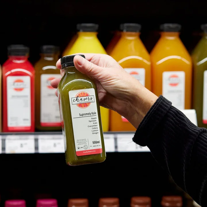 A person holding a bottle of Chasers Fresh Juic green juice labeled "Supremely Kale" in a store aisle with other juice bottles in various colors behind it.