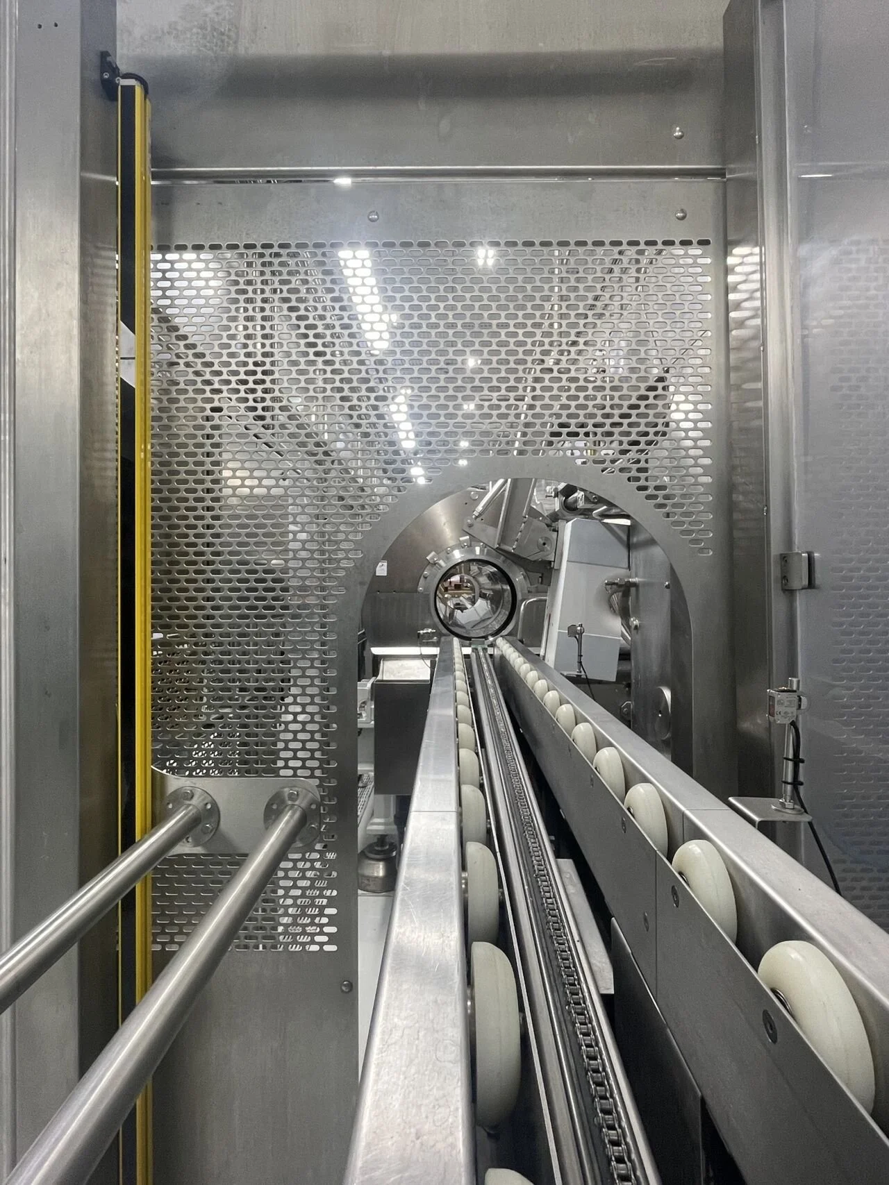 Industrial HPP machine with a conveyor belt and rollers, enclosed in a stainless steel box with perforated panels.