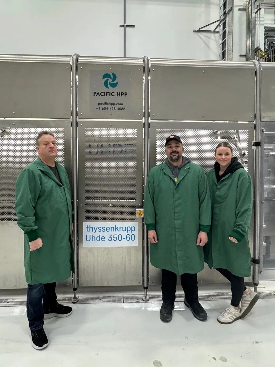 Three workers in green lab coats standing in front of industrial equipment with signs that reads 'PACIFIC HPP'