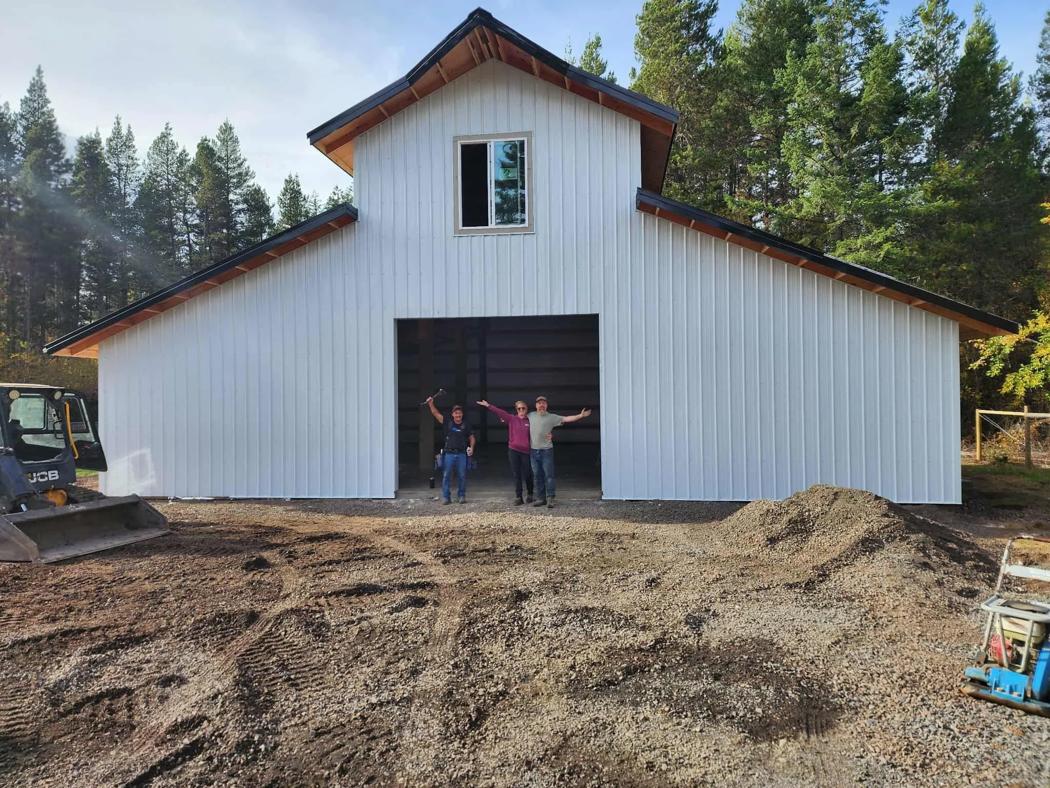Three people standing in the entrance of a new white barn with a large open doorway during daytime. One person is holding a hammer, celebrating, with trees in the background.