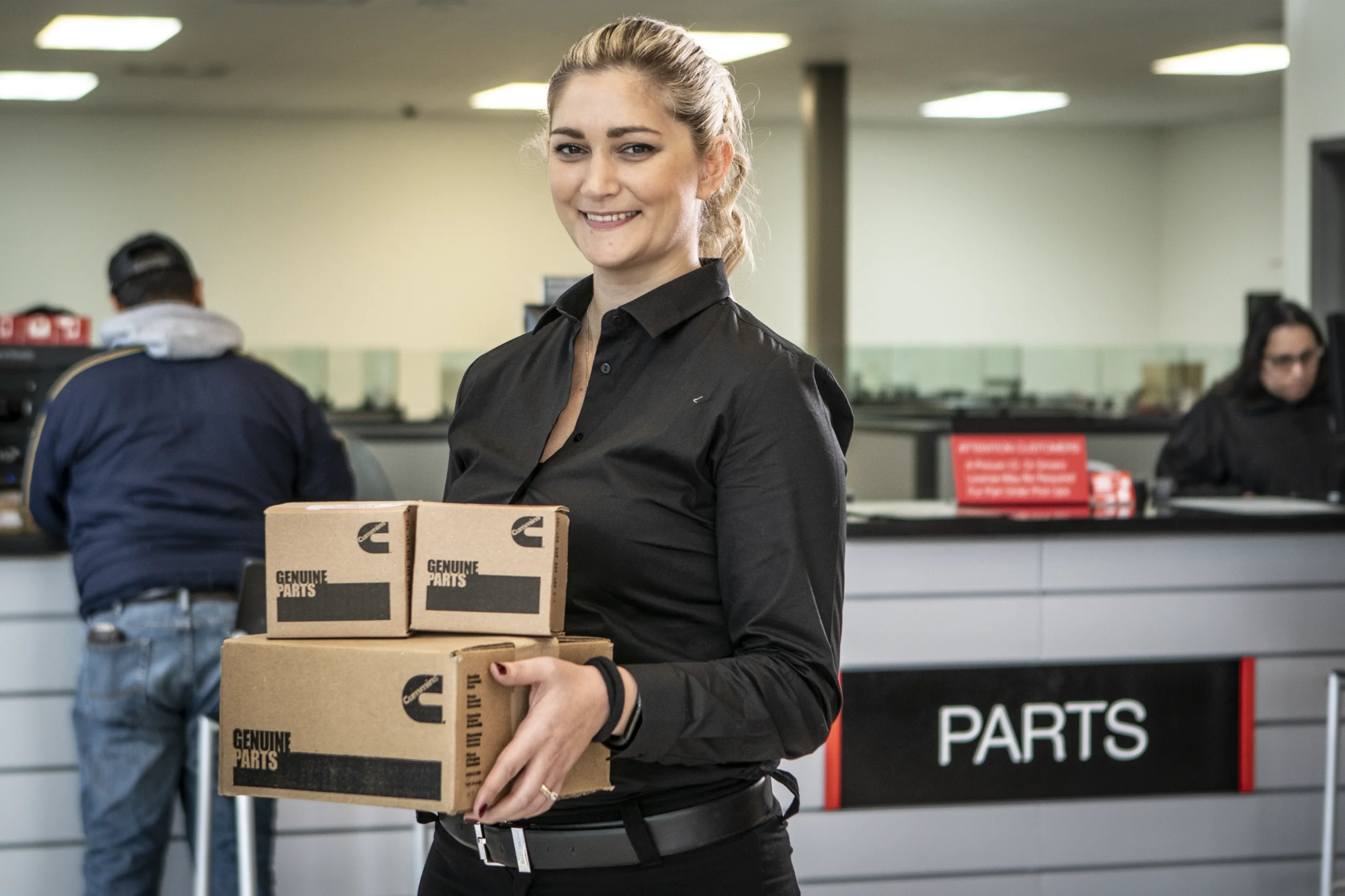 Corporate lifestyle portrait of a female employee holding parts boxes inside a professional service department, photographed for branding and recruitment.